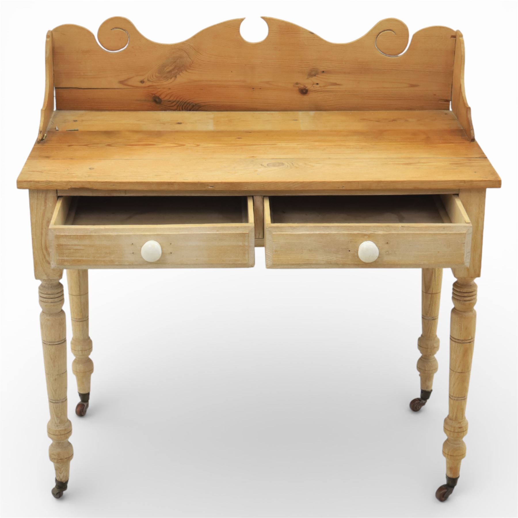19th century stripped pine side table, the rectangular plank top with low shaped back rail incorporating scroll cut ends, fitted with two frieze drawers with ceramic knob handles, raised on turned supports terminating in castors