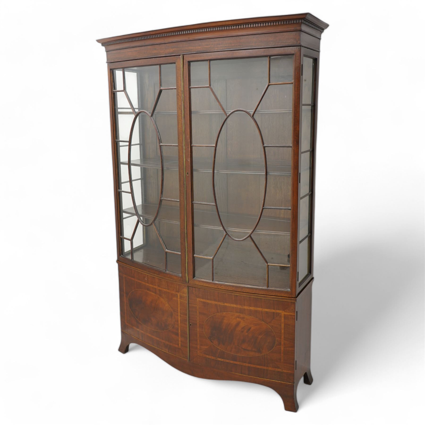 Edwardian mahogany and satinwood bow-fronted display cabinet, moulded dentil cornice over two astragal glazed doors, fitted with three shelves, double cupboard below, the doors inlaid with figured oval panels and satinwood bands, shaped apron on splayed bracket feet 