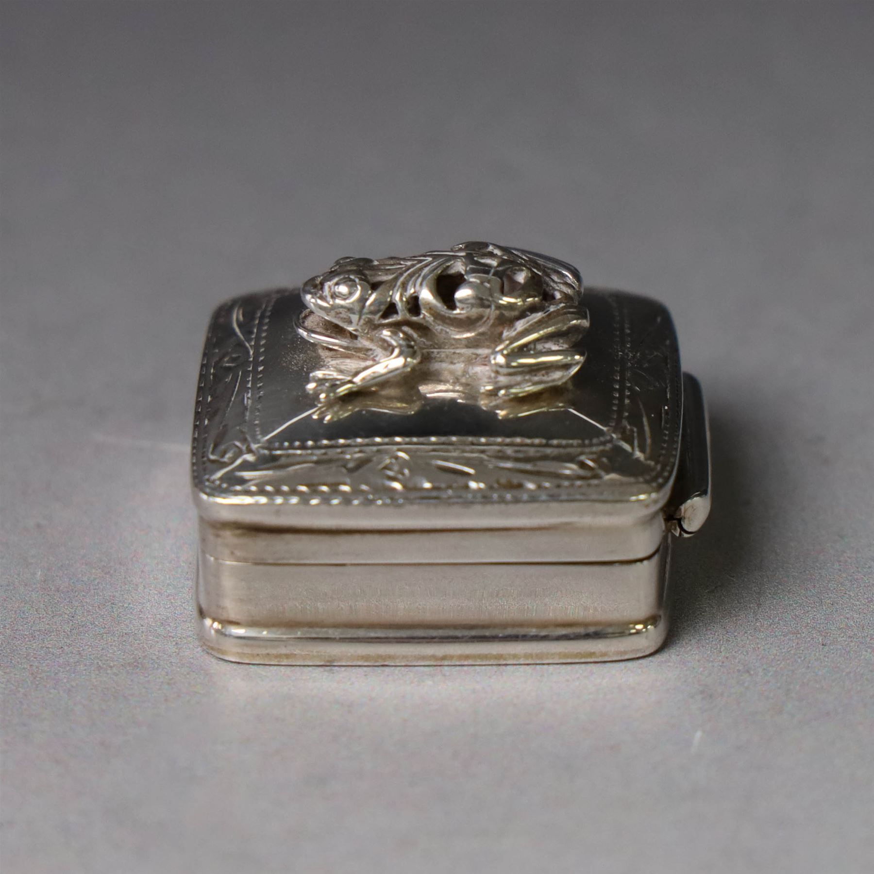 Modern silver pill box, of square form, the hinged cover surmounted with figure of a frog within a engraved scroll edge, hallmarked hallmarked Martin Gerald & Sid Adler, London 2000, W3cm