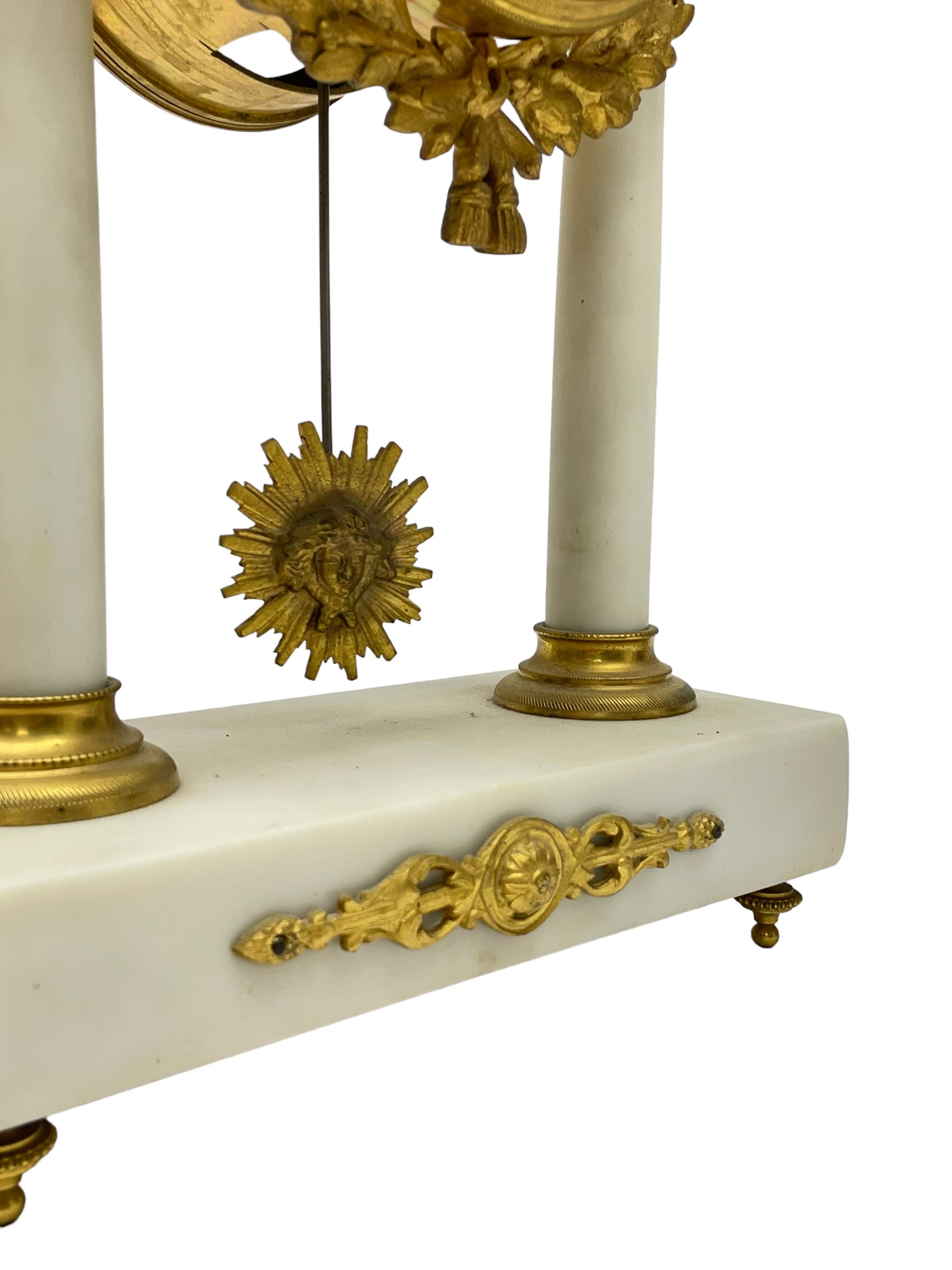 French - Late 19th century 8-day white marble and gilt metal portico clock garniture, the clock surmounted by an urn and raised on a pair of cylindrical pillars above a rectangular plinth base, white enamel dial with Arabic numerals and floral garlands, twin train countwheel striking movement, striking the hours and half hours on a bell, with a sunburst pendulum and gilt Louis XVI hands, garniture with matching twin light candelabra. With Key.   Candelabra H 30cm.