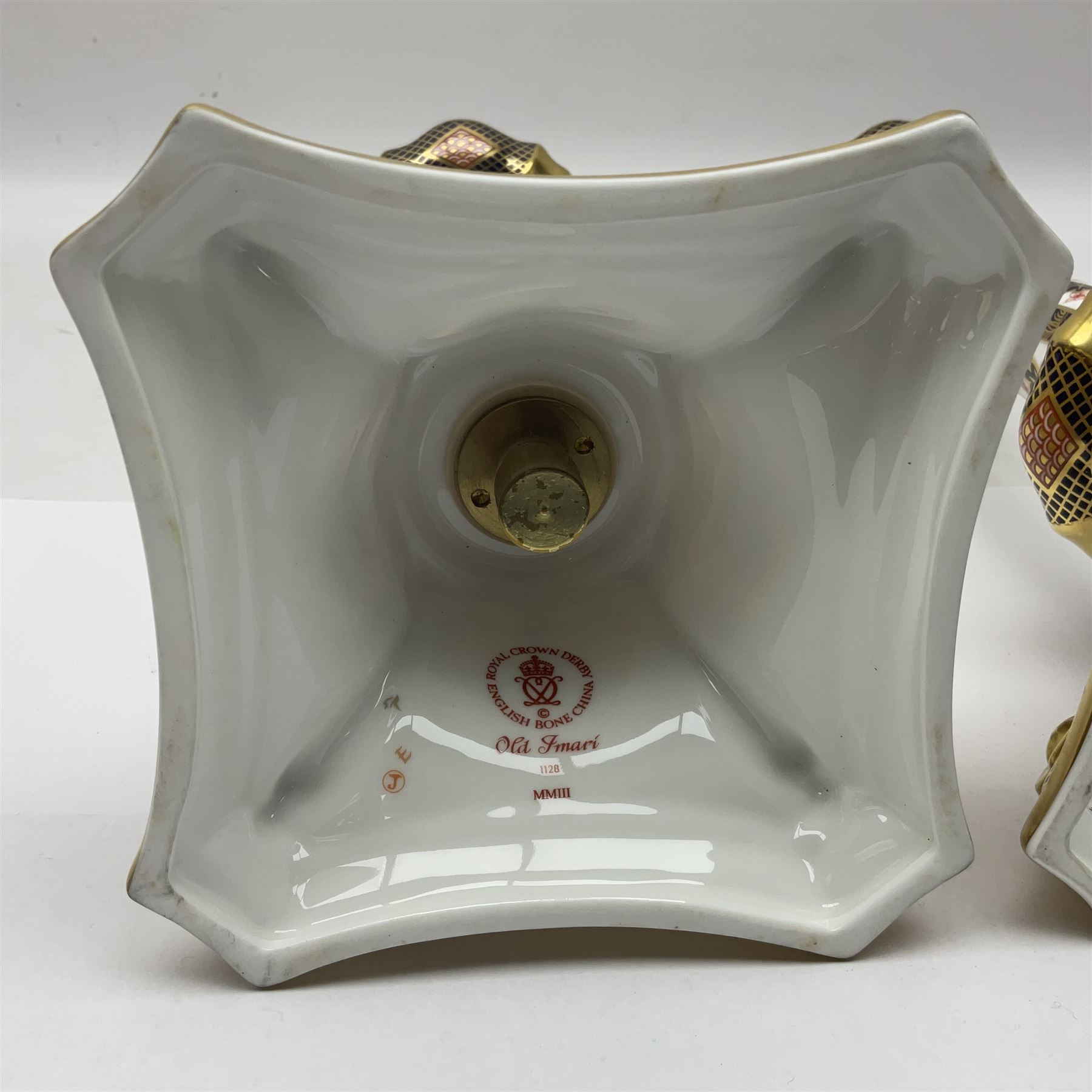 Pair of modern Royal Crown Derby Old Imari 1128 pattern candlesticks, each with shaped drip tray affixed to squat socket, upon spreading stem and canted square base with gilt dolphin edges, with printed marks beneath including Roman numeral year cypher for 2003, H26.5cm, with original boxes