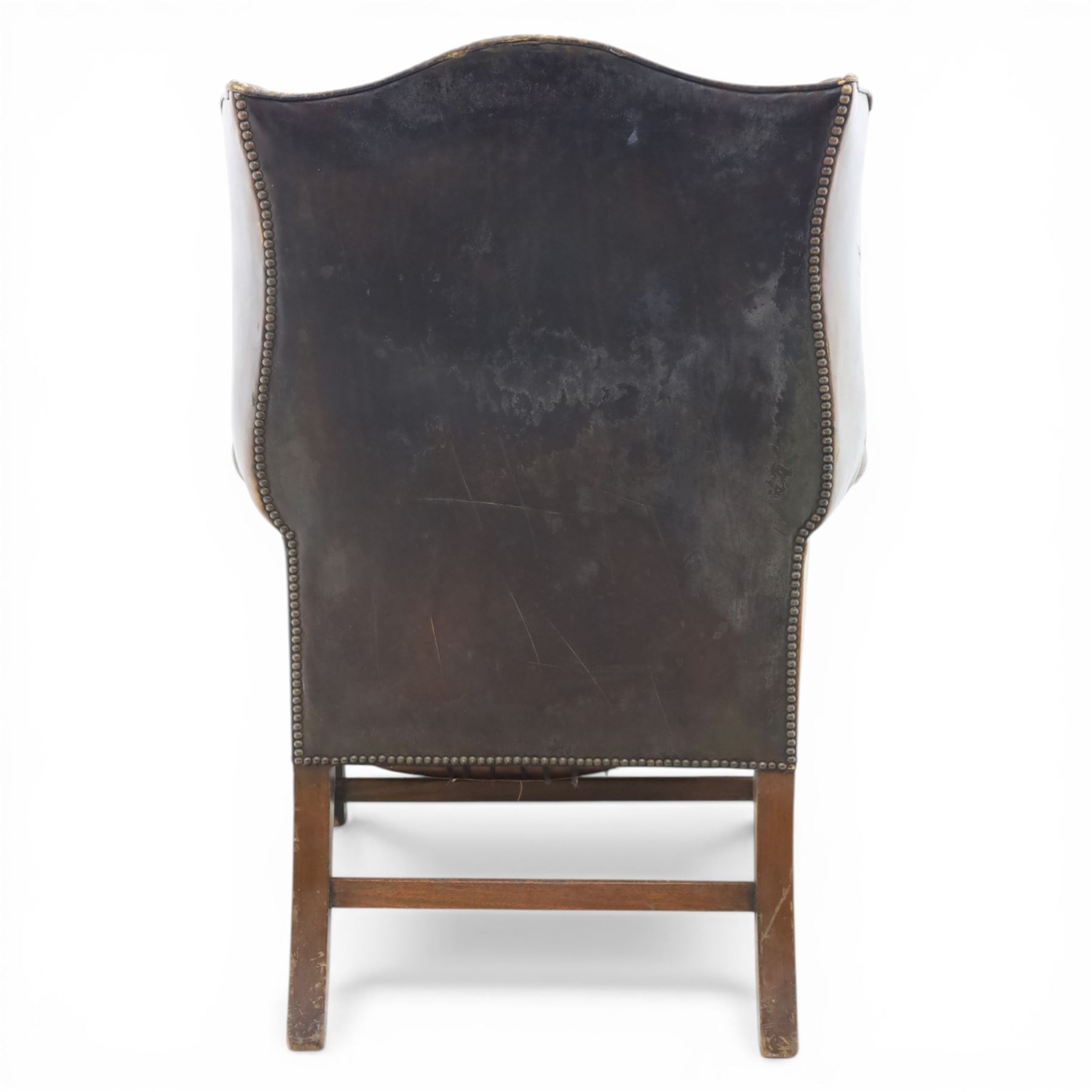 Georgian style leather wing armchair, shaped high back with outscrolled wings above padded arms and deep buttoned seat, close-nailed throughout, raised on square supports united by H-stretcher