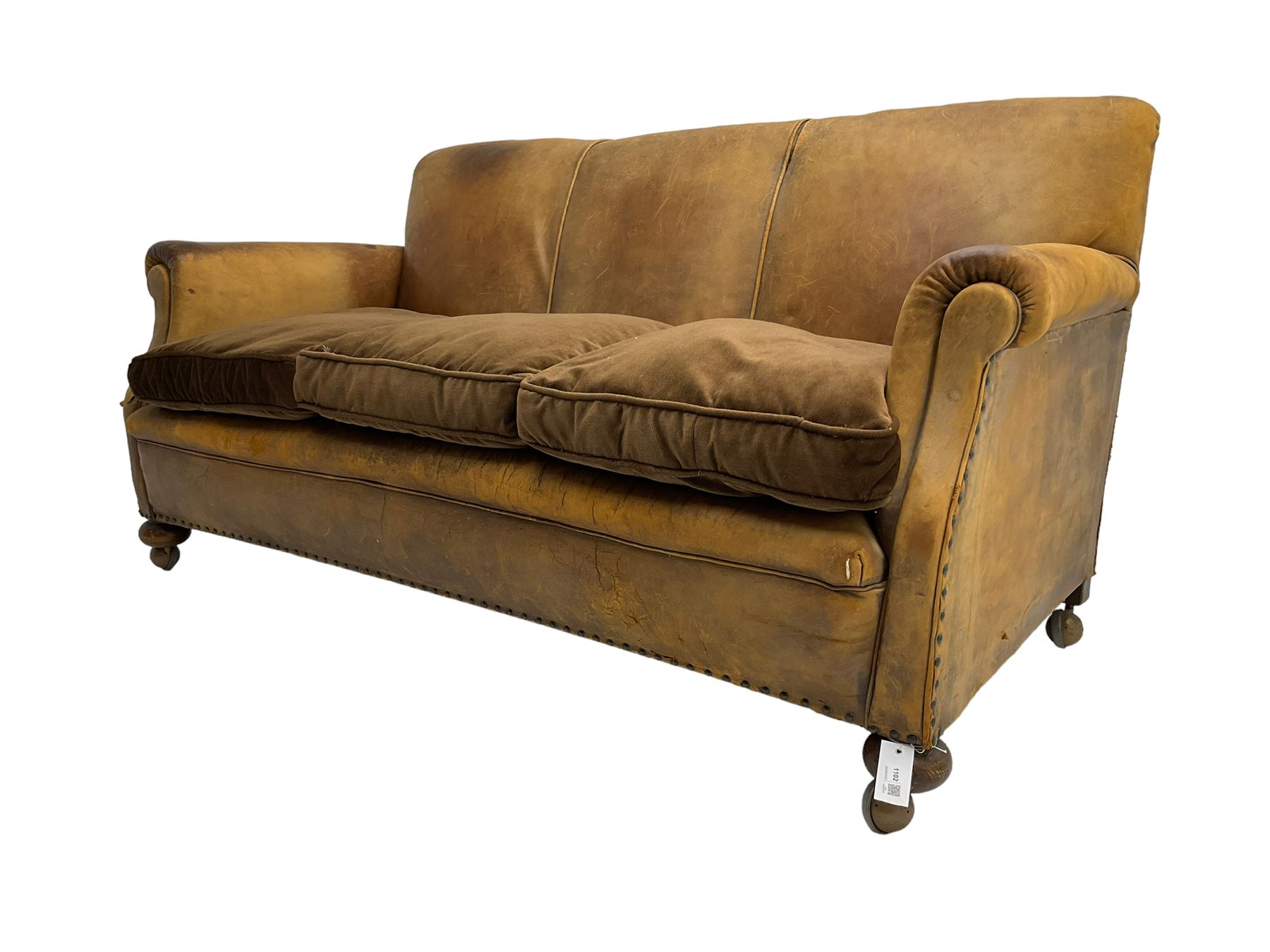 Early-to-mid-20th century three seat sofa, upholstered in worn tan leather with studwork, loose cushions upholstered in brown fabric, on compressed bun feet with castors