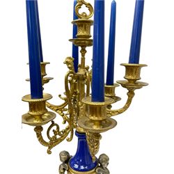 A 20th century continental gilt metal Lyre mantle clock with a pair of matching six light candelabra, spring driven movement housed in a gilt drum case on a brass and cobalt blue base with paw feet, two bronzed metal figures of mythological cherubs with animal legs and cloven hooves supporting a festooned garland swag, eight-day twin barrel striking movement with a floating lever balance escapement, striking the hours and half-hours on two bells, white enamel dial with roman numerals, minute track and pierced steel hands, dial inscribed “Imperial”. 