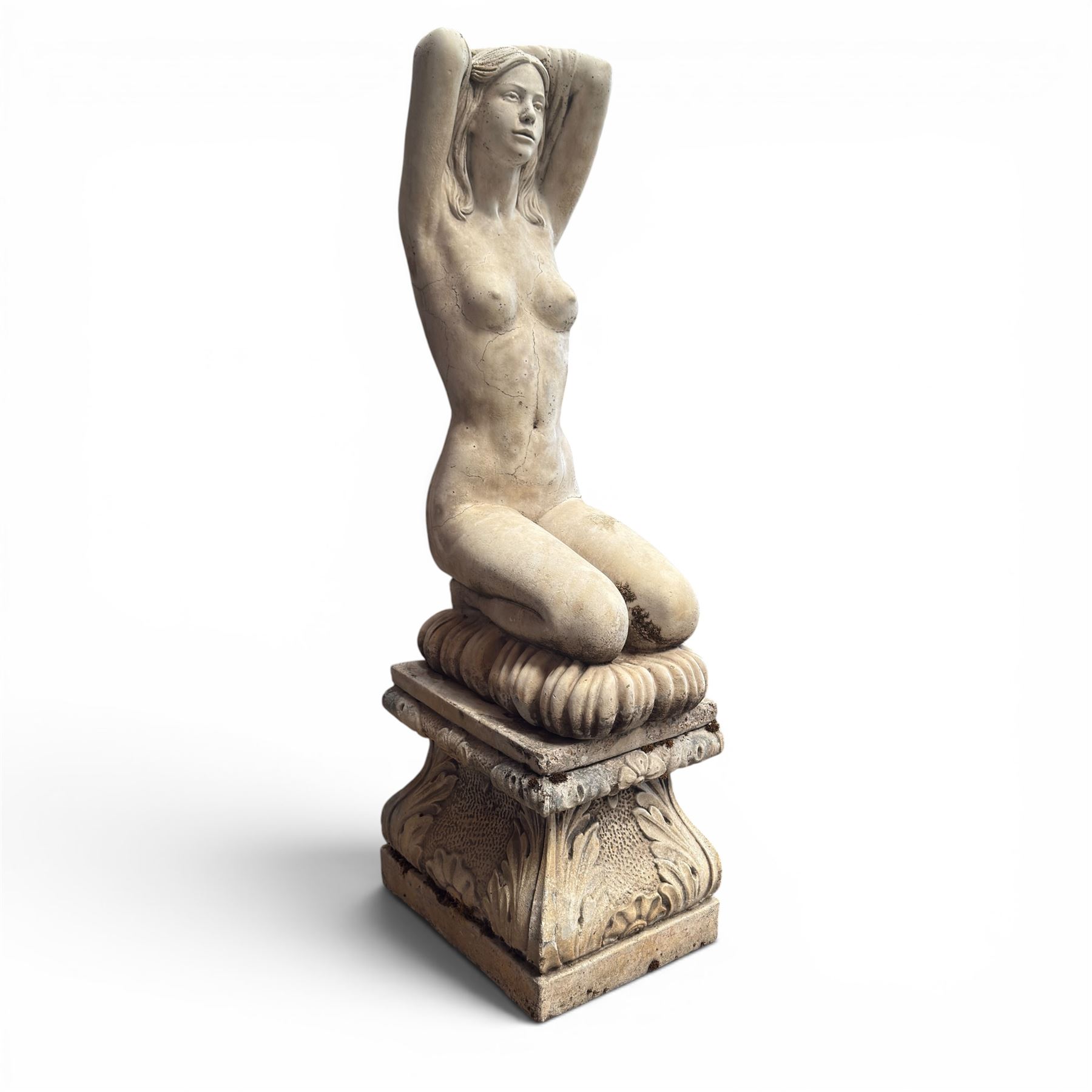 20th century composite stone garden figure, modelled as a kneeling nude female with arms raised behind her head, mounted on an integral cushion, the squared ogee pedestal moulded with acanthus leaves and flower heads, in a white painted and weathered finish