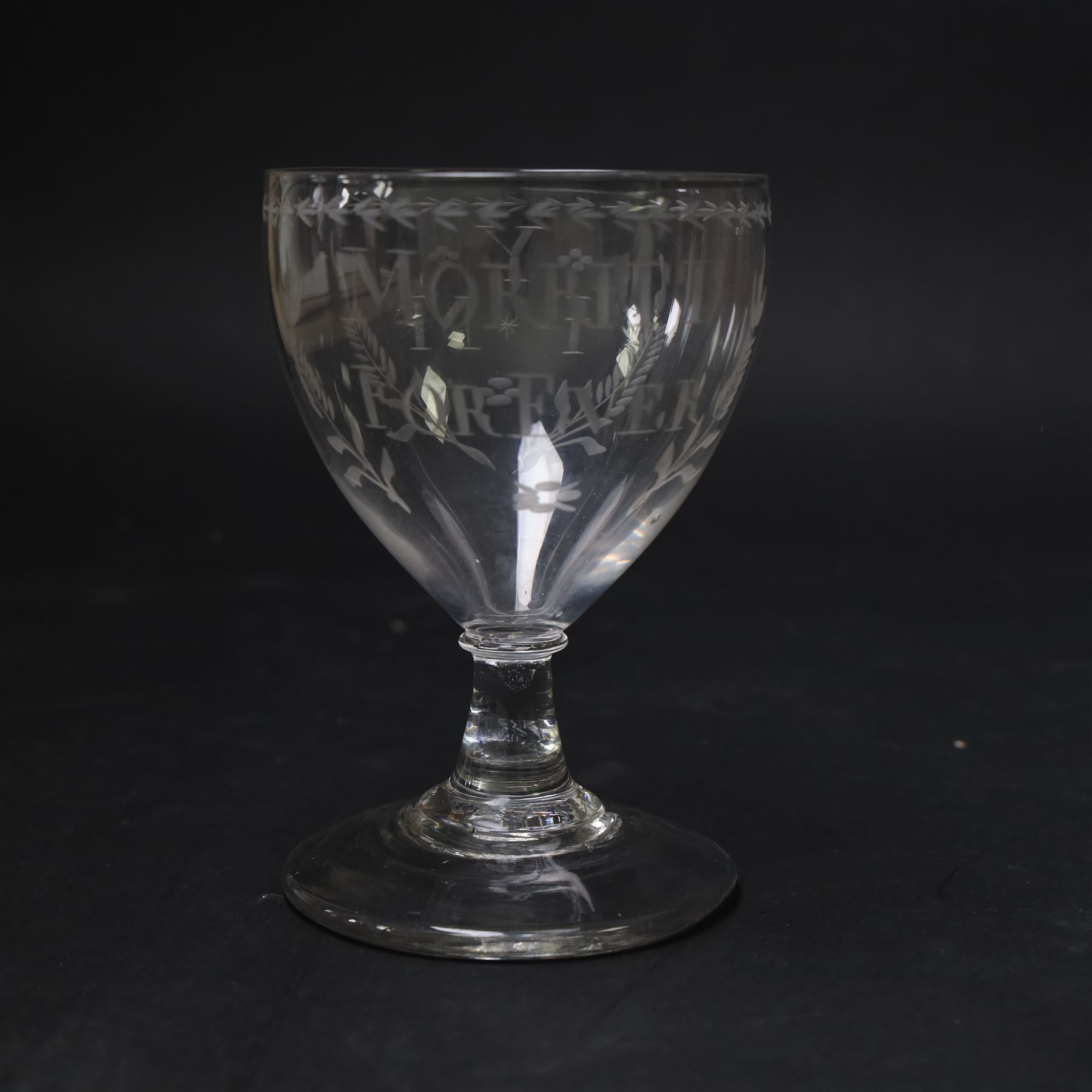 Early 19th century electioneering glass rummer, circa 1800-1830, the funnel bowl engraved with the inscription 'MORRITT FOREVER', and initials 'Y I M' verso, within leafy sprigs and beneath a husk band to rim, upon a plain short spreading stem, upon conical foot, H13.5cm

John Bacon Sawrey Morritt (circa 1772–1843) of Rokeby Park was a traveller, classical scholar, landowner, politician, and friend of Sir Walter Scott. He sat in Parliament for Beverley (1799–1802) and later for Northallerton (1814–1818), periods when fiercely contested elections often produced engraved drinking glasses as campaign propaganda, which were distributed in taverns and at election dinners. Examples connected to local candidates such as this are particularly scarce and survive as rare intersections of political history and material culture. 