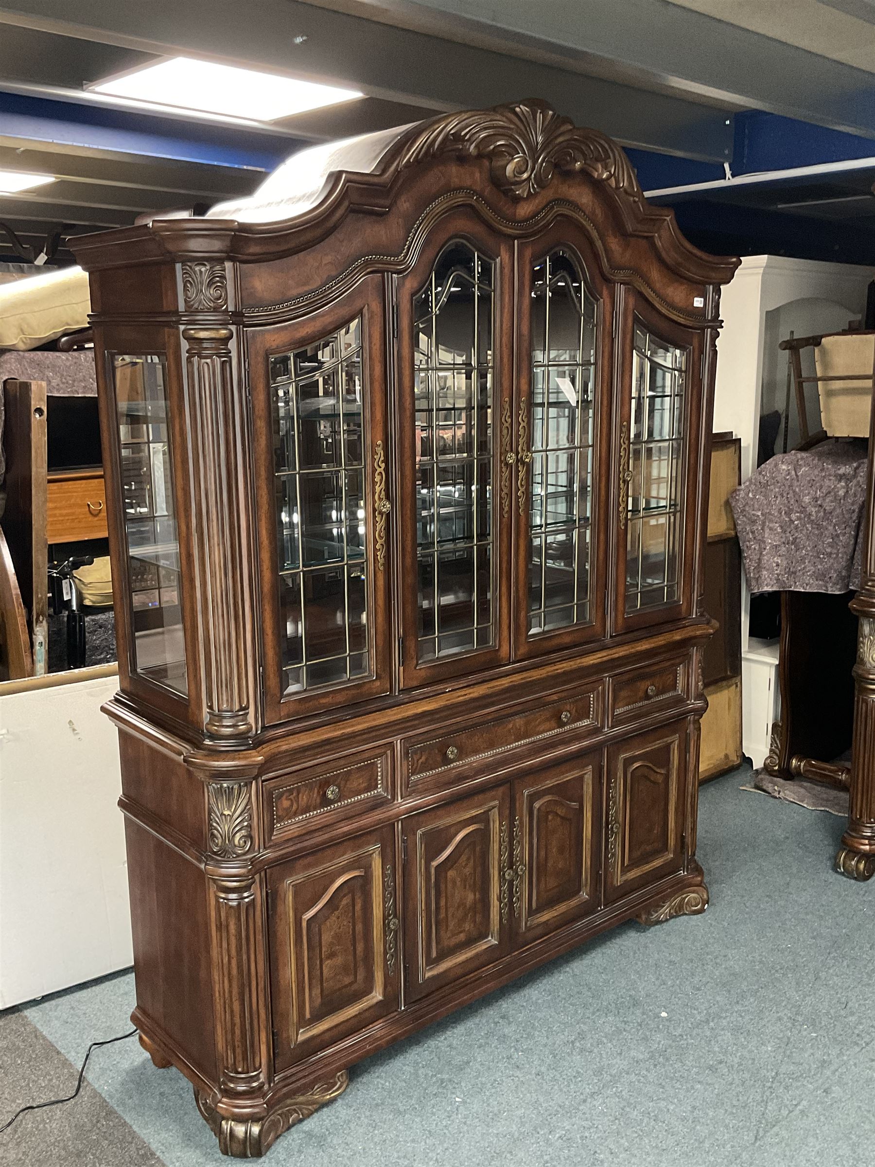 Kevin Charles American walnut display cabinet, illuminated interior, shell carved pediment above four bevel edge doors enclosing six glazed shelves above one central long drawer flanked by two short drawers, above three cupboards, carved bracket supports 
