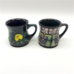 Two Moorcroft mugs of waisted form, the first example decorated in the Violet pattern designed by Sally Tuffin, the second example decorated in the Buttercup pattern designed by Sally Tuffin, each with impressed marks beneath, H8.5cm. 