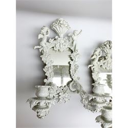 Set of four late 19th/early 20th century Continental girandole white glazed wall sconces, the shaped mirror backs within foliate detailed frames surmounted by putti, supporting two scrolling and floral encrusted branches above ribbon terminals, with blue underglaze mark to sides, H38cm
