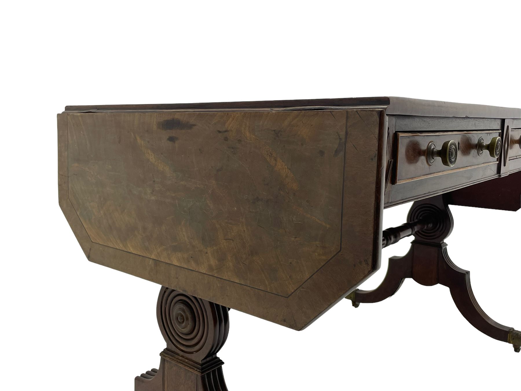 Regency mahogany sofa table, canted rectangular drop leaf top with walnut crossbanding, fitted with two frieze drawers with central lozenge decoration, two opposing false drawers, vasiform end supports with reeded edge and central roundel joined by ring turned and reeded stretcher, splayed supports on scaled brass cups and castors