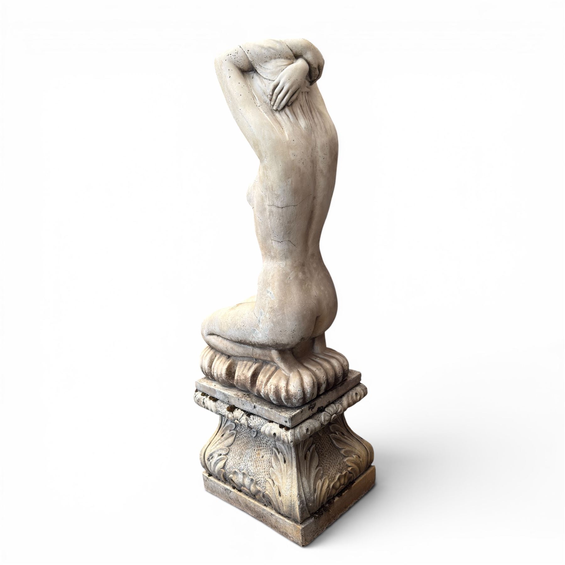 20th century composite stone garden figure, modelled as a kneeling nude female with arms raised behind her head, mounted on an integral cushion, the squared ogee pedestal moulded with acanthus leaves and flower heads, in a white painted and weathered finish