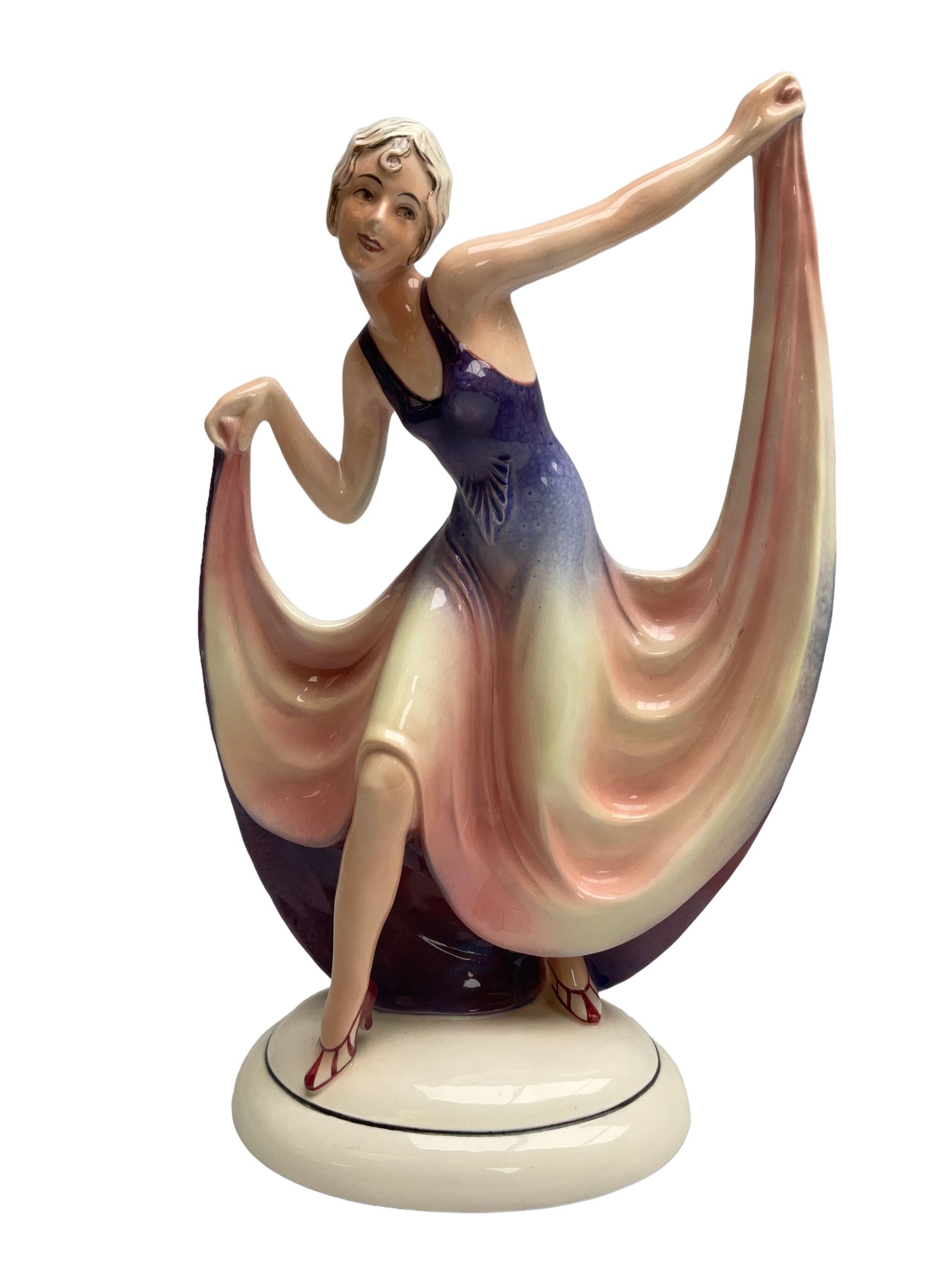 Early 20th century Hertwig Katzhütte figure of an Art Deco lady dancing, wearing a pink and purple dress, stood upon on a domed plinth, with green cat H house printed mark beneath, H32cm