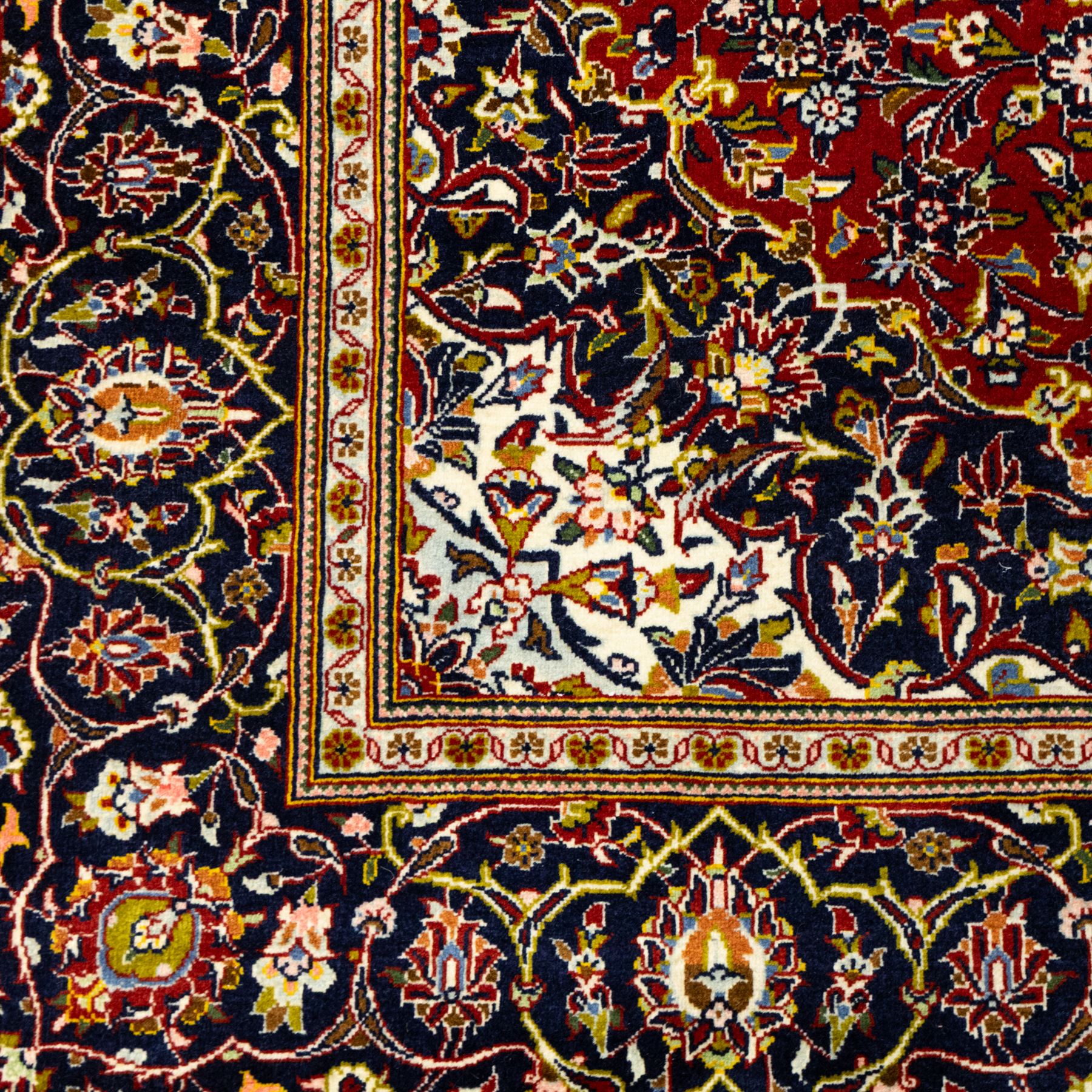 Central Persian Kashan hand-knotted carpet, crimson ground, with a central ivory shaped floral medallion with pendants, surrounded by dense scrolling foliate and floral motifs incorporating rosettes, palmettes and delicate vine work, enclosed by ivory spandrels and a dark navy border with repeating palmettes and meandering vines, flanked by floral guard stripes, with cotton fringes