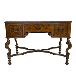 20th century figured walnut writing table, rectangular top with inset leather writing surface and foliate carved edge, fitted with three drawers over kneehole with S-scroll carved brackets, on acanthus carved S-scroll supports terminating to turned feet, united by a shaped and stepped X-framed stretcher rails with moulding 