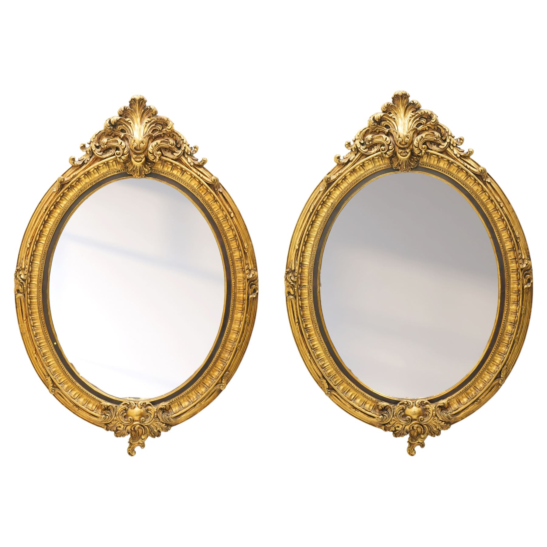 Pair of Rococo Revival gilt framed oval wall mirrors, the exuberant pediment formed of scrolling acanthus leaves and rocaille ornament, the oval bevelled plate set within with ebonised slip with outer repeating lappet decoration, over a rocaille cartouche terminal