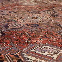 North West Persian Heriz hand-knotted carpet, madder red field with central stepped geometric medallion with radiating angular motifs and serrated outlines, and stylized floral and tribal devices framed by a broad border of repeating angular palmettes and rosettes flanked by multiple guard stripes, with cotton fringe ends 