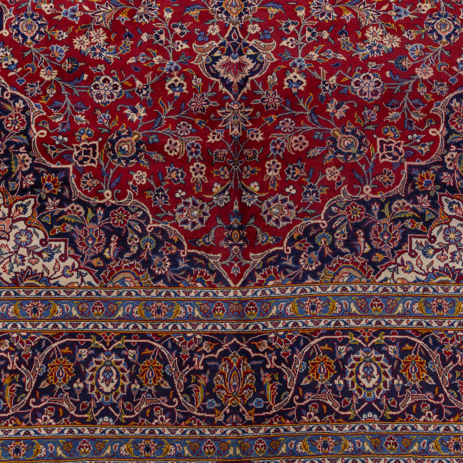 Central Persian Kashan hand-knotted carpet, madder red field with central cusped medallion in navy and ivory enclosing a floral rosette and radiating scrolling arabesques and palmettes, across a densely ornamented ground of finely drawn vinery enriched with a profusion of flowering sprays and stylized blossoms, spandrels in deep indigo and principal border of navy ground decorated with a continuous palmette and vine-scroll flanked by multiple guard stripes, with cotton fringe ends
