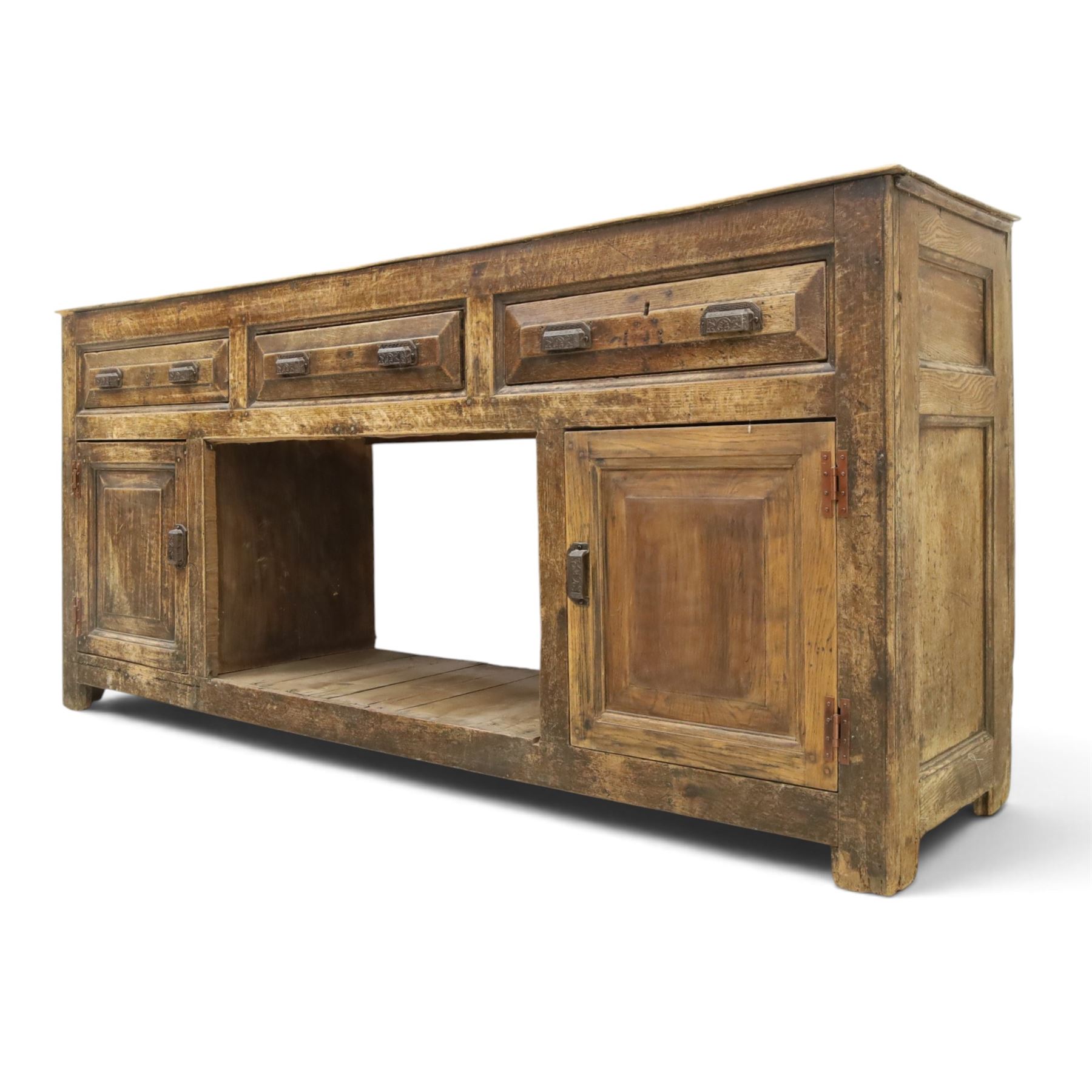 18th century rustic oak dresser base, with fielded panels and iron cup handles with embossed greek key and foliate detail, three short drawers above and open recess flanked by two cupboard doors, raised on stile supports