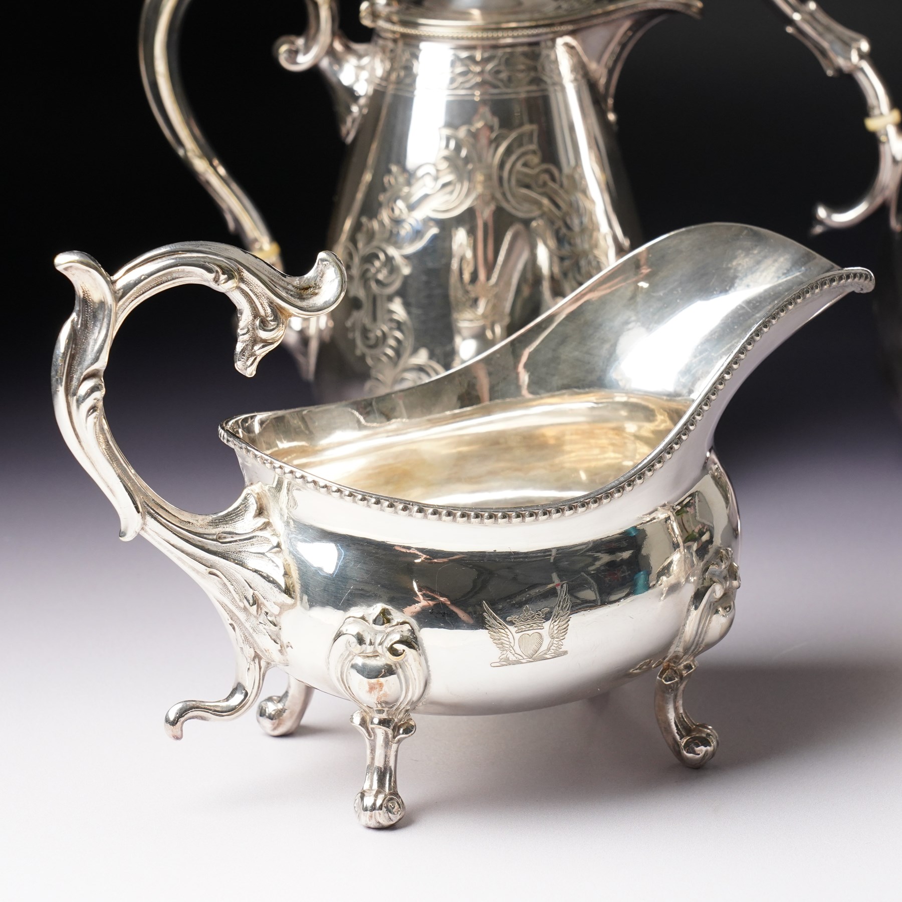 Pair of large silver plated sauce boats with bead edge decoration and C scroll handles engraved with a crest in the form of a winged heart and on shaped supports L20cm Maker probably Hawksworth Eyre & Co and an engraved hot water jug and a matching smaller jug with the same crest and by the same maker