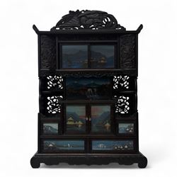 Early 20th century Japanese Meiji style carved hardwood cabinet, of symmetrical tiered form with pierced foliate fretwork panels and phoenix crest, fitted with sliding and hinged doors inset with hand-painted landscape panels depicting Mount Fuji, waterfalls, and lakeside dwellings, raised on shaped bracket supports