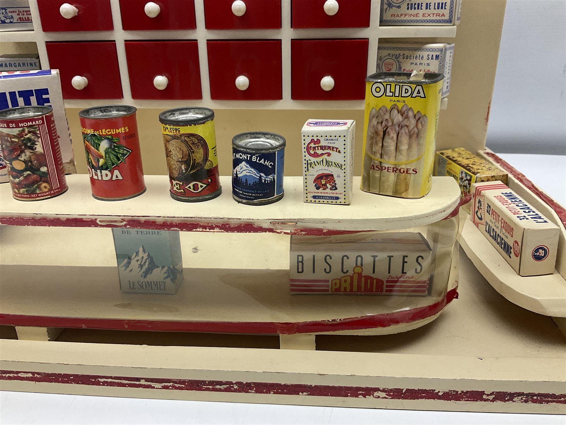 Late 1950s continental red and white painted wooden diorama of a grocery shop interior with fitted shelves and drawers and freestanding 'glazed' counter, fully stocked with miniature tin cans and packets of food L42cm H23cm D22cm
