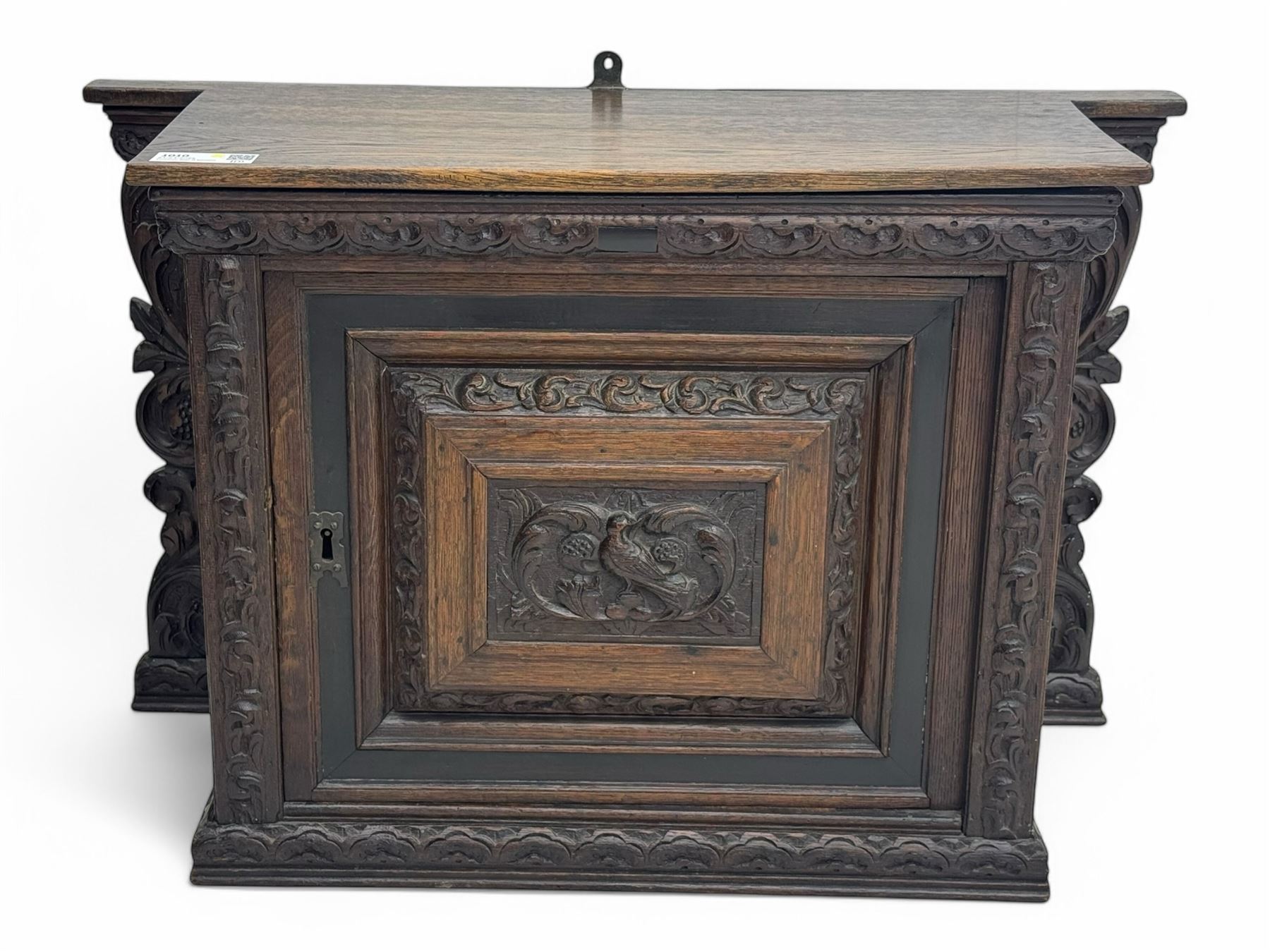 18th century and later carved oak cupboard, enclosed by panelled door carved with central bird motif and scrolled leafage with an outer ebony band, the frieze and uprights carved with repeating lunette and floral designs, flanked by scrolled foliate carved upright brackets