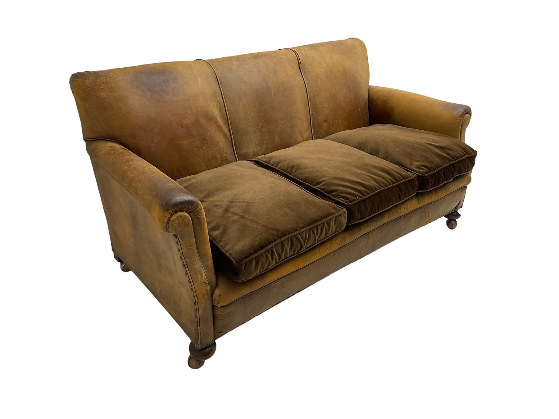 Early-to-mid-20th century three seat sofa, upholstered in worn tan leather with studwork, loose cushions upholstered in brown fabric, on compressed bun feet with castors