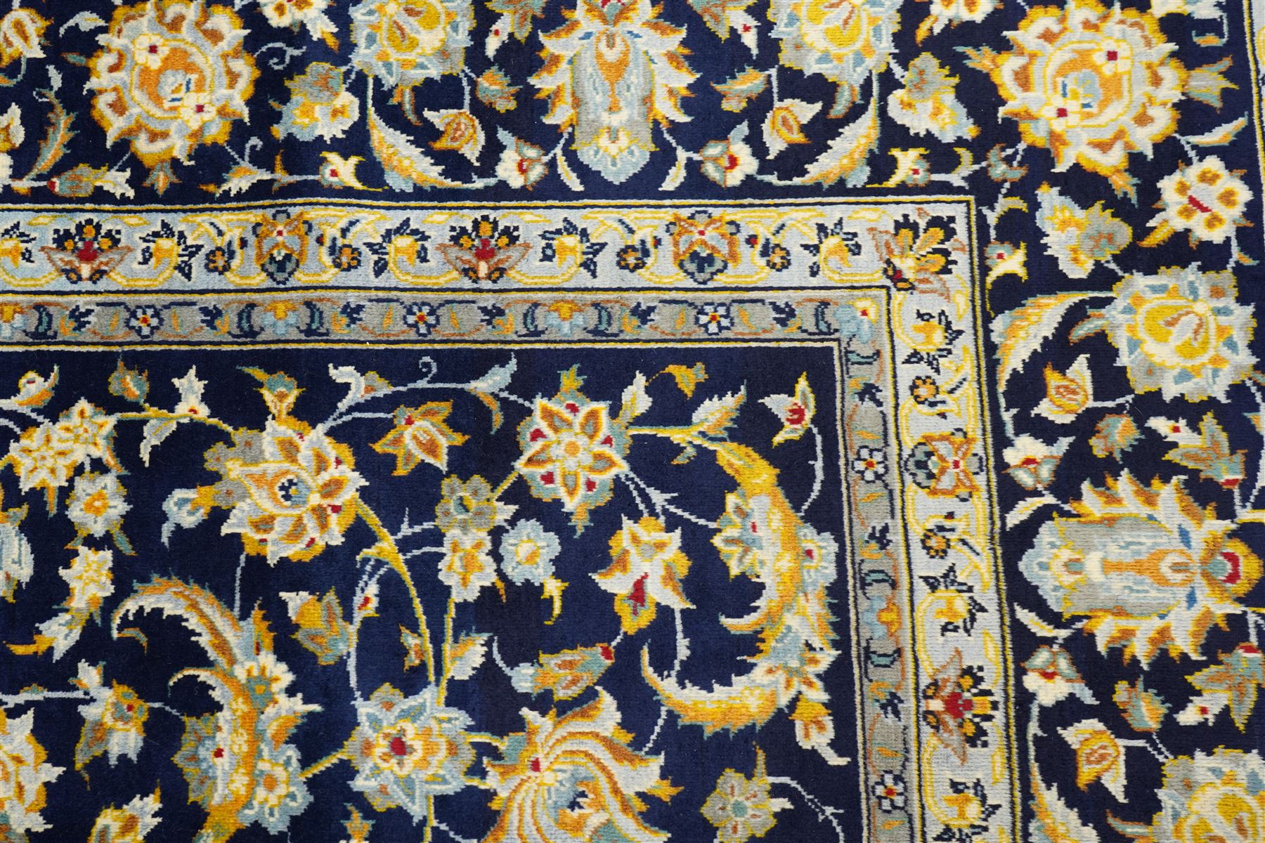 Central Persian Kashan hand-knotted carpet, deep indigo field with a densely all-over arrangement of scrolling floral vines, palmettes, and flowering sprays, in soft ivory, pale blue, and warm gold tones, principal ivory border with a continuous meandering vine of stylized palmettes, and floral motifs, flanked by multiple narrow guard borders, with cotton fringe ends 