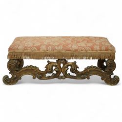 19th-century Rococo-style giltwood window seat, upholstered in floral-patterned coral-ground fabric with fringed border, the intricate scrolled supports united by pierced scrolling foliate stretchers with acanthus and C-scroll decoration