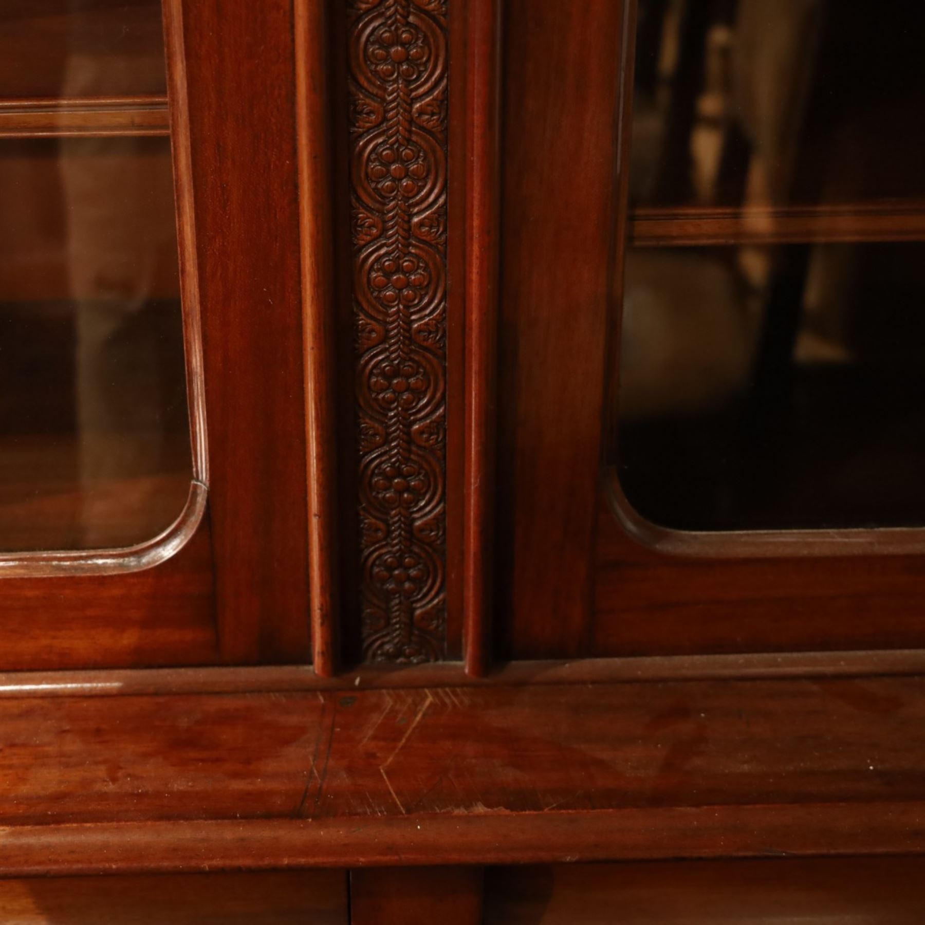 Victorian carved mahogany enclosed bookcase on cupboard, projecting moulded cornice over twin glazed doors enclosing adjustable shelves, flanked by foliate carved uprights, the lower section fitted with two cushion moulded drawers over twin panelled cupboard doors, divided and flanked by conforming carved uprights, on plinth base
