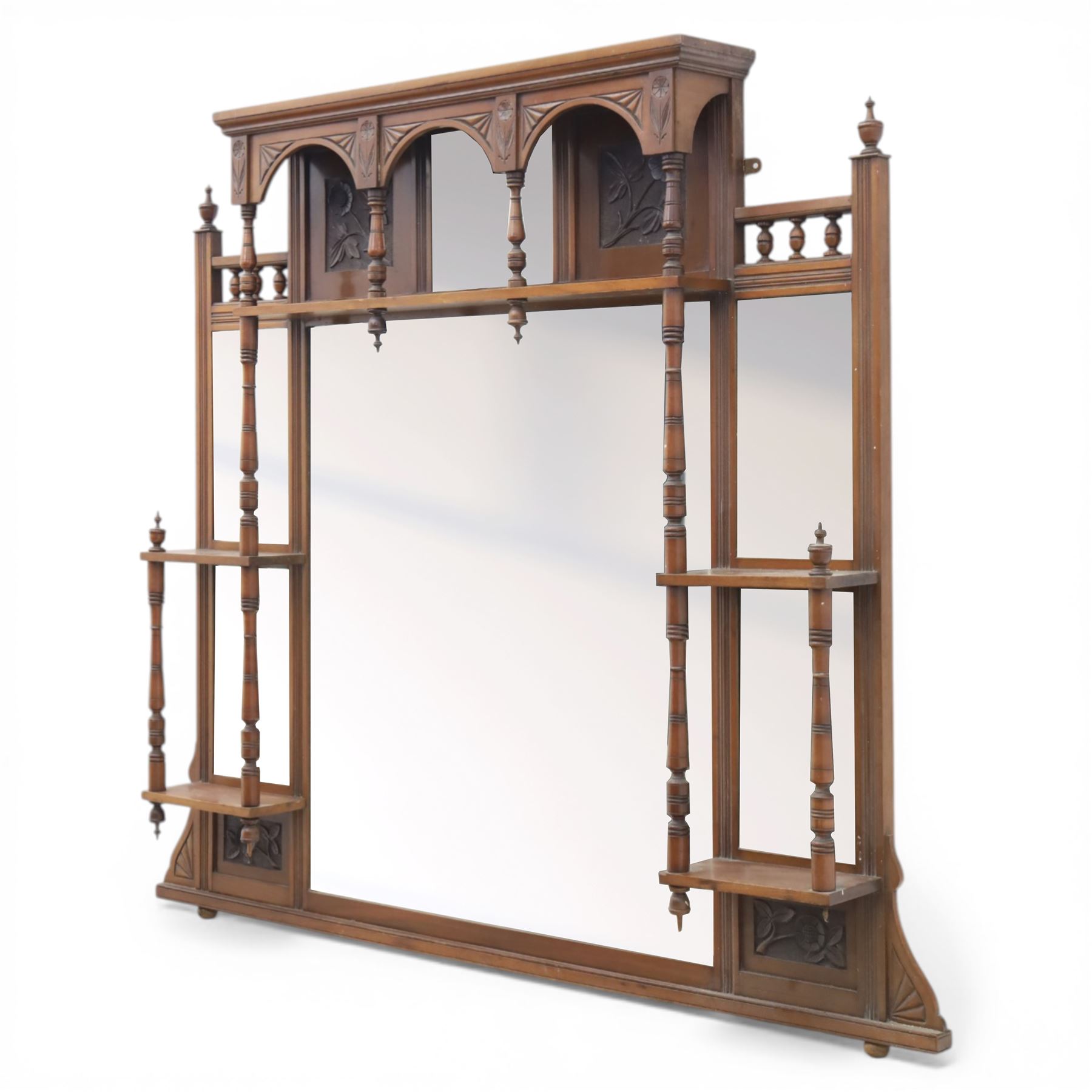 Edwardian walnut overmantel mirror, moulded cornice over three arched apertures with turned supports, flanked by open shelves with turned uprights and finials, carved foliate panels to the base