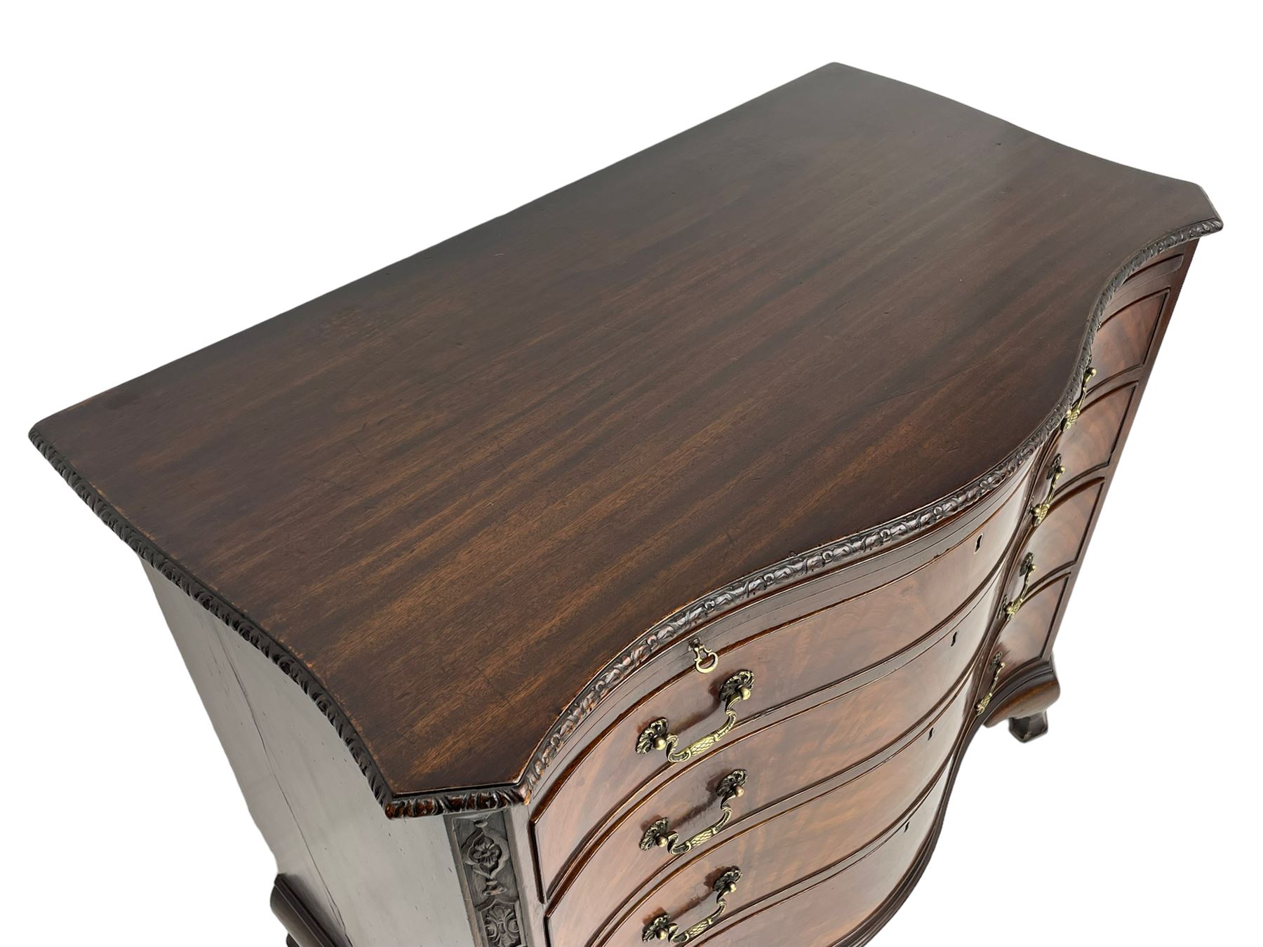 Chippendale design serpentine chest, shaped moulded top carved with repeating flower head motifs, fitted with brushing slide over four graduating drawers, pierced and foliate moulded handle plates with scrolled swan neck handles, canted upright corners carved with flower heads and strapwork, on canted ogee bracket feet