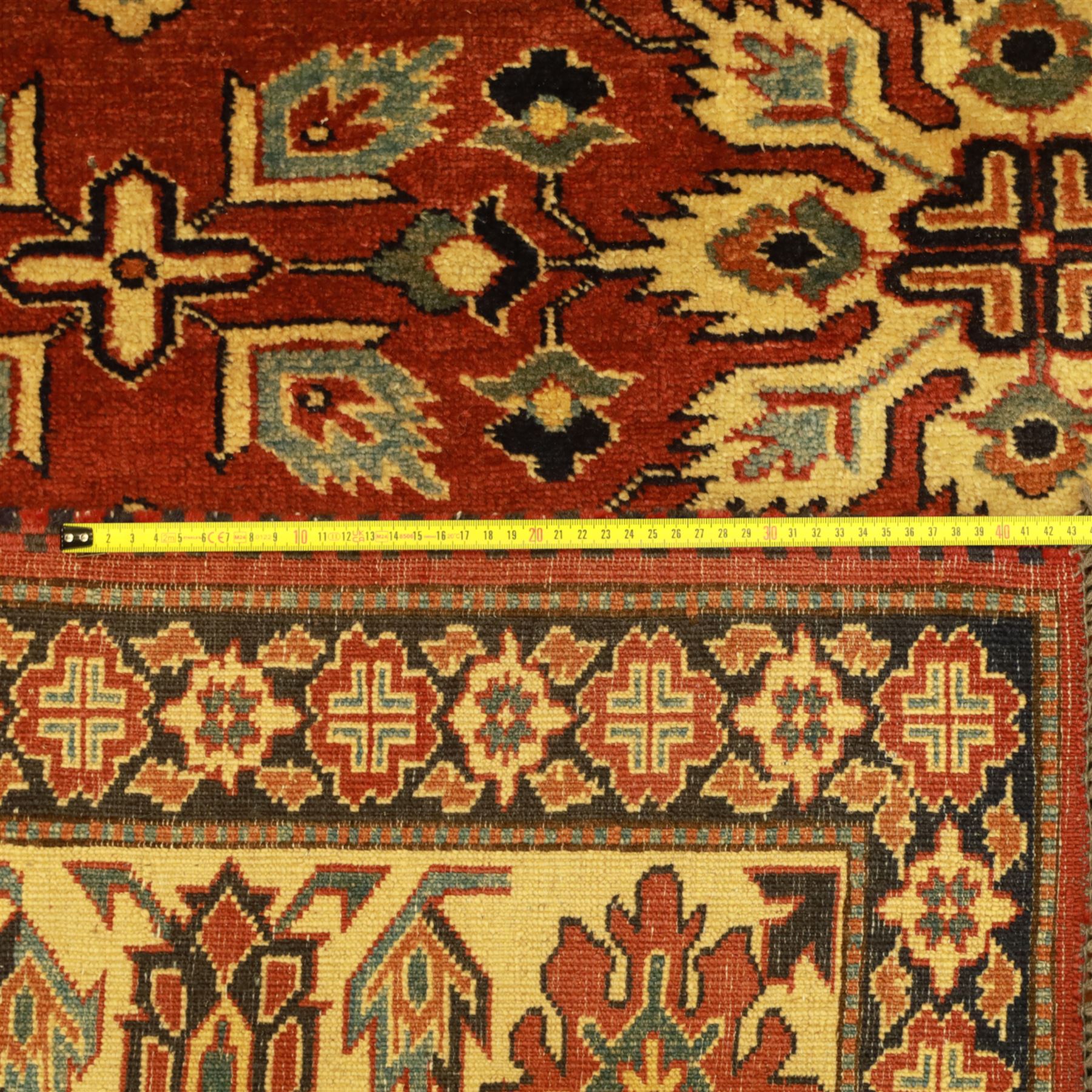 Afghan Gazni Kazak rug, the hand-knotted wool pile featuring a rust red ground with all-over geometric floral motifs in ivory and dark green, enclosed by a wide ivory border with stylised rosettes and angular vine decoration, fringed ends, from the region of Ghazni, Afghanistan