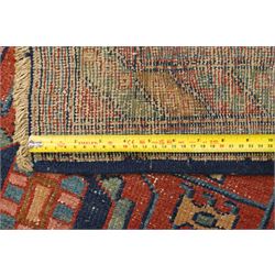 Persian Heriz hand-knotted carpet, terracotta ground centred by large dark navy geometric medallion with stepped outline, decorated throughout with stylised flowerheads, palmettes and geometric motifs in blue, green and ochre, enclosed by diagonal geometric border with narrow guard stripes