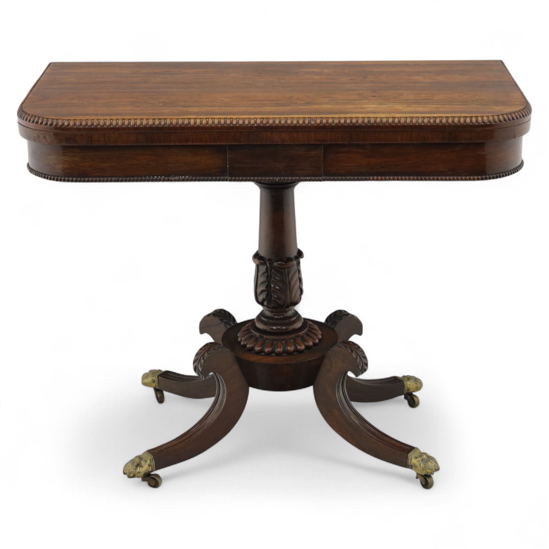 Regency rosewood fold over card table, the rectangular top with rounded corners and gadrooned edge opening to reveal a green baize lined interior, raised upon convex lobbed and acanthus pedestal and four fluted sabre legs, terminating in brass lion paw feet and castors