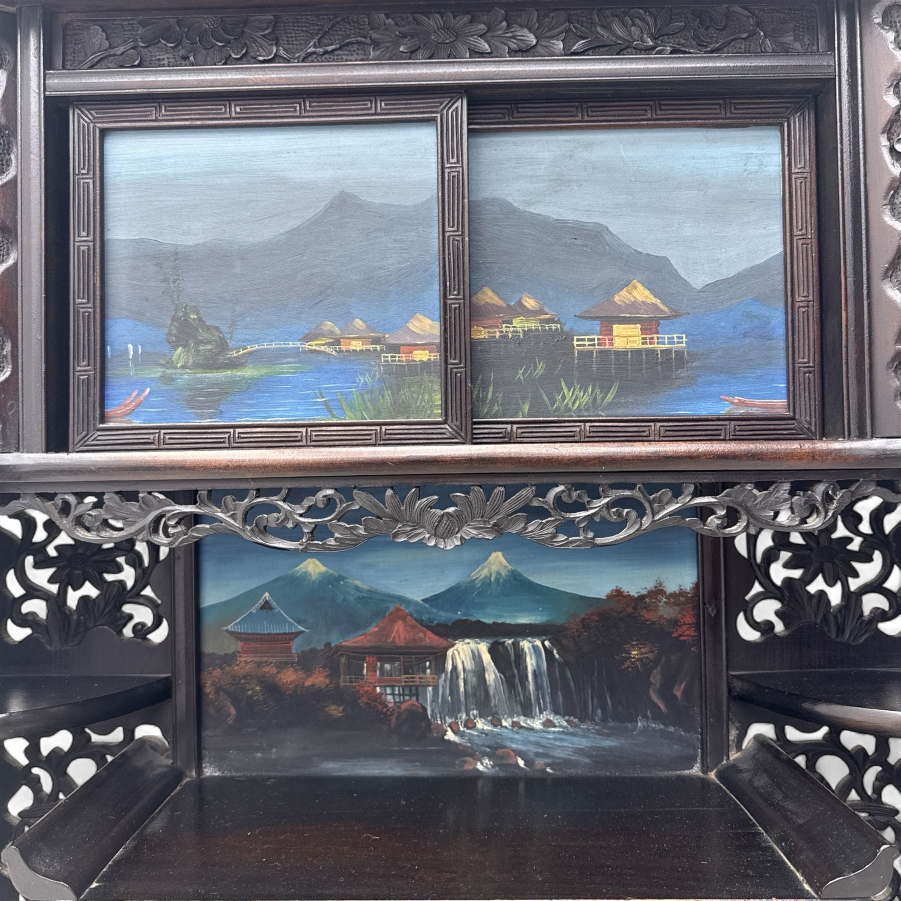 Early 20th century Japanese Meiji style carved hardwood cabinet, of symmetrical tiered form with pierced foliate fretwork panels and phoenix crest, fitted with sliding and hinged doors inset with hand-painted landscape panels depicting Mount Fuji, waterfalls, and lakeside dwellings, raised on shaped bracket supports