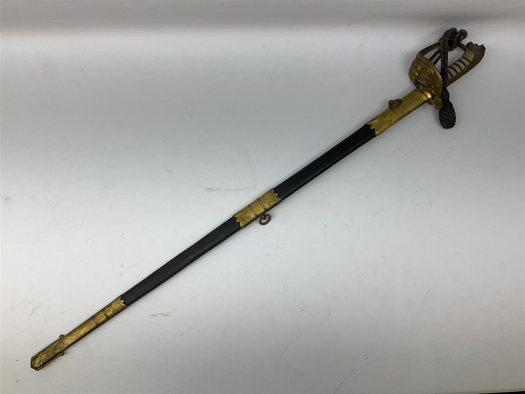 WW1 George V era 1827 Pattern Royal Naval Officer’s sword, the 80cm fullered steel blade decorated with crown, fouled anchor etc and marked Gieves Portsmouth London Edinburgh Devonport to the ricasso, gilded hilt with fouled anchor and crown, wire-bound ray skin grip, lion’s head pommel with mane backstrap, folding/locking langet and full bullion knot; in  leather scabbard having twin brass lockets with hand engraved panels and twin suspension rings L94cm overall. Provenance: By direct family descent from Brian Heathcote HMS Conqueror later Superintendant in the Liverpool Special Constabulary.