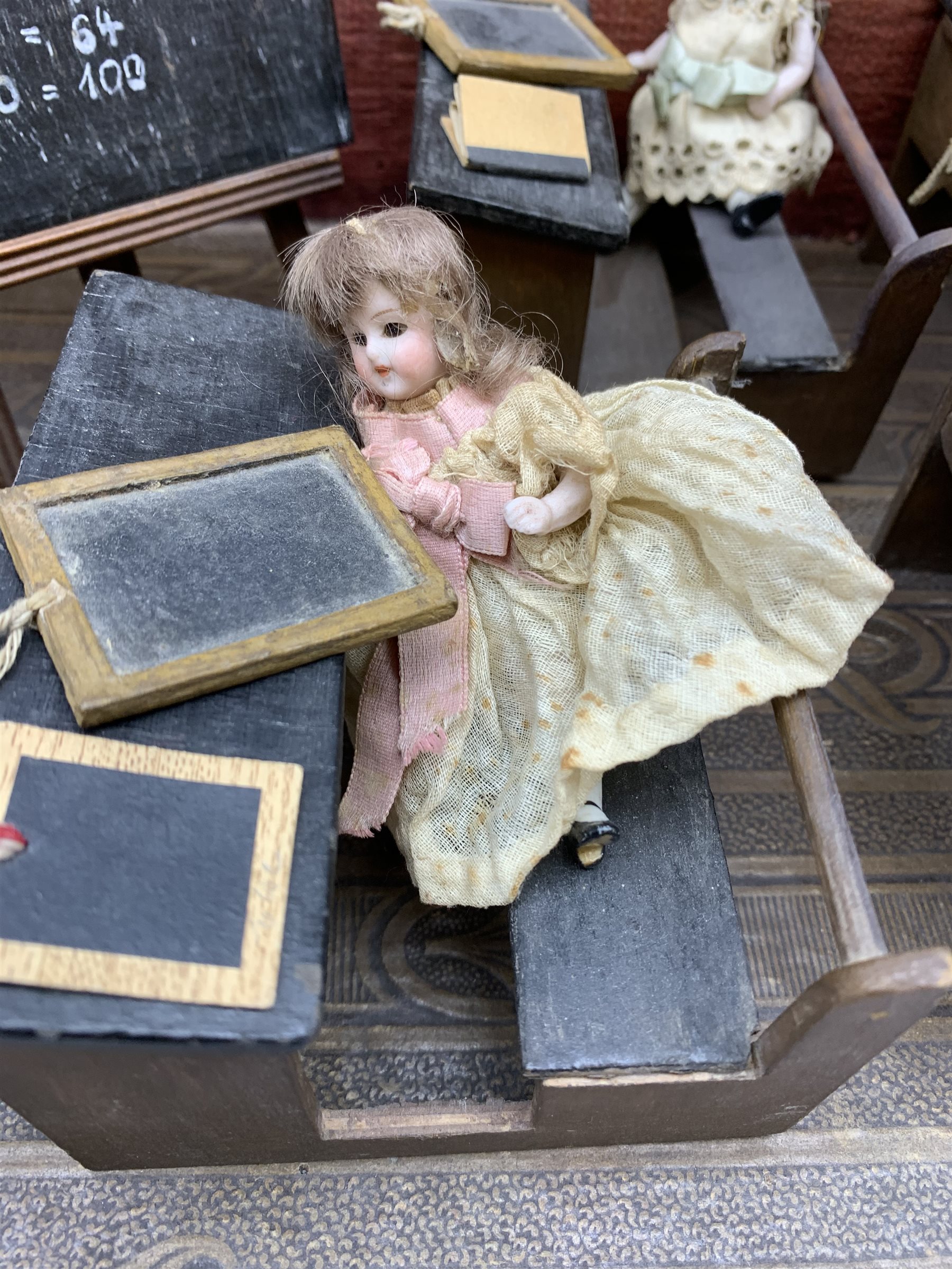 Late Victorian wooden school room setting with bisque head teacher and six German all bisque pupils each seated at a combined desk and chair with slate board, in a classroom furnished with chest of drawers, fire-surround, lectern, wall map, pictures, miniature books, school bell, chalk board on easel, wall clock etc, L70cm H28cm D28cm
