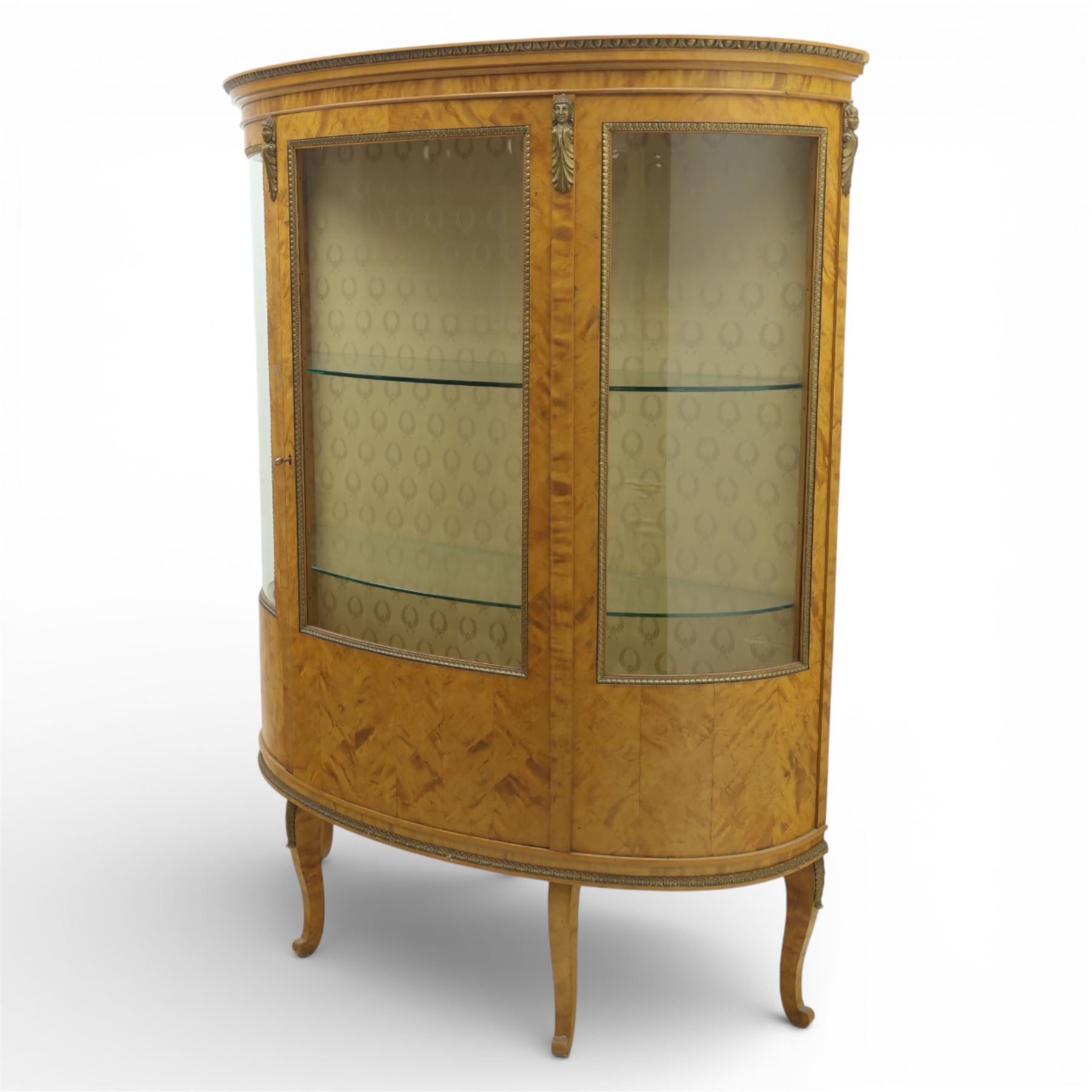 Edwardian satin walnut display cabinet, bowed oval form with moulded and dentil cornice over glazed panels, enclosing a laurel wreath damask interior with adjustable glass shelves, the frame applied with gilt metal bead mouldings and mask mounts, enclosing a shelved interior, raised on cabriole supports with moulded feet