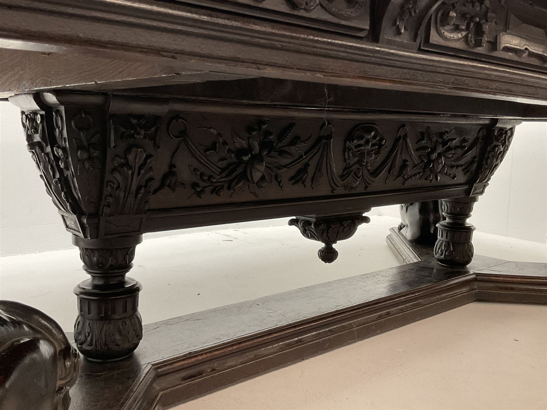 Large 19th century carved oak library table, the rectangular top with lunette carved edge over cartouche panelled and carved  scroll frieze, the central tapering rectangular support relief carved with masks and garlands, on four carved seated lion supports at each corner, on flat angular stretcher with recessed castors stamped ‘Bartholomew & Fletcher, London’