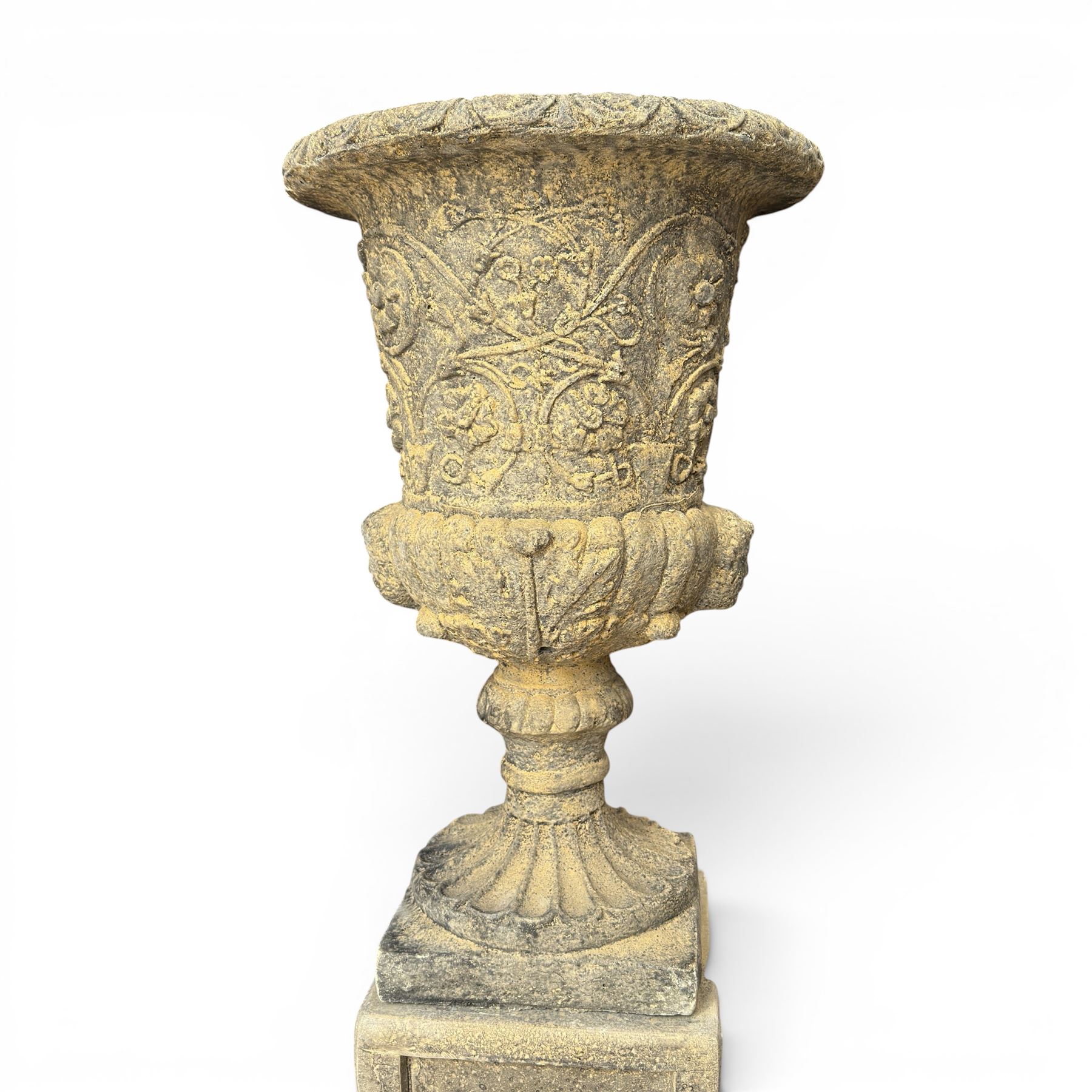 Pair of George III design antiqued composite stone garden urns of campana form, the egg-and-dart everted rim over a waisted body relief moulded with scrolling fruiting foliage and flowerheads, the gadrooned underbelly with twin mask ornaments, the knopped stems on fluted socles, raised on faux panelled square pedestal bases