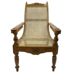 Late 20th century teak framed plantation chair, cresting rail and apron carved with two elephants with upraised trunks, flanked by finials, cane back and seat, fitted with hinged extending leg rests, on ring turned supports