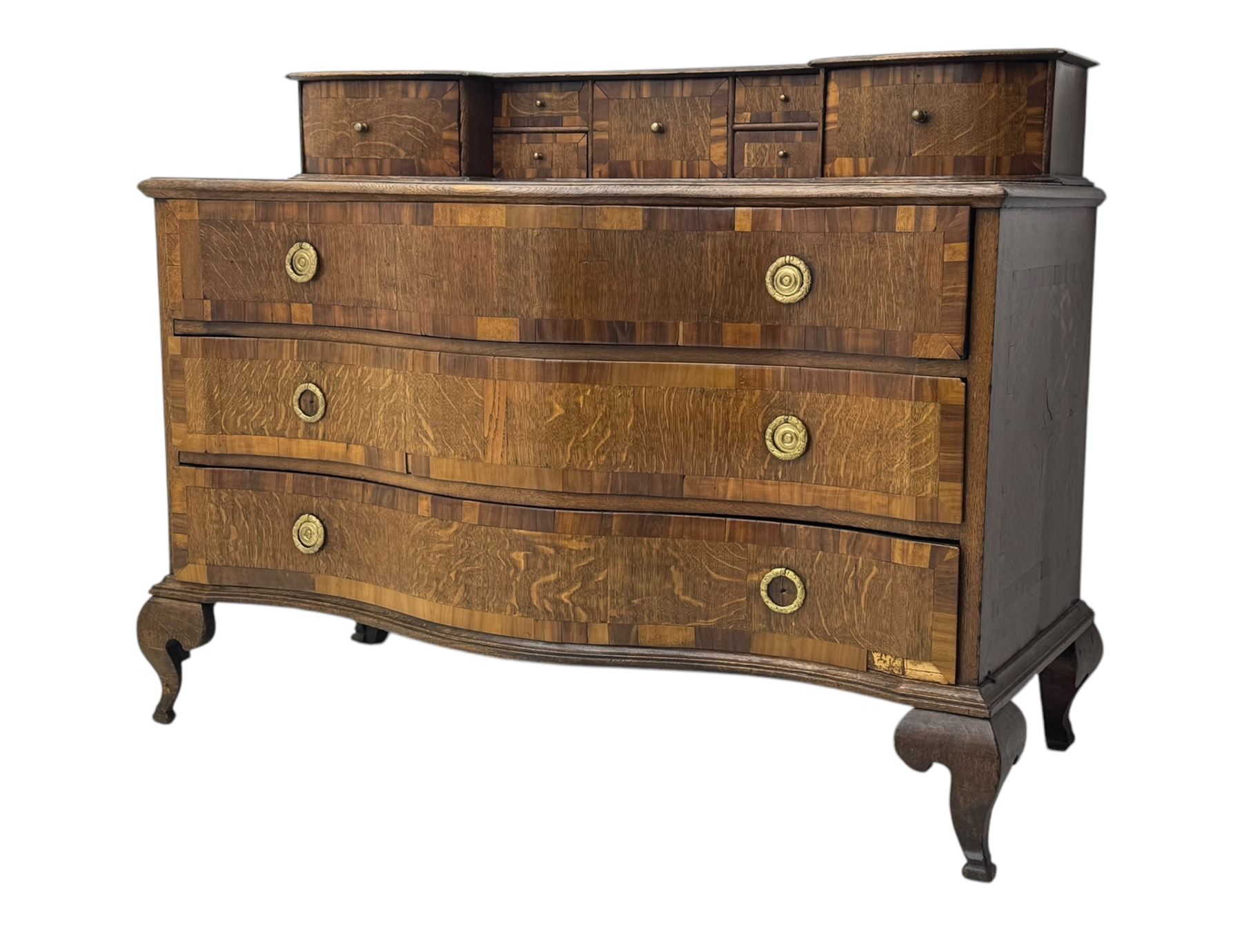 Late 19th century continental oak commode, serpentine fronted with parquetry and crossbanded decoration, the raised superstructure back fitted with a central bank of five recessed drawers flanked by concave drawers, above three full-width drawers with brass ring handles, raised on cabriole supports