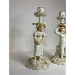 Pair of Royal Worcester figural candlesticks designed by James Hadley in the style of Kate Greenaway, modelled with a young boy and girl with painted features beside stylised ivy clad tree stumps supporting naturalistic sconces and drip pans, raised upon rocky circular bases, decorated with gilt detail throughout, with printed puce stamped marks and impressed 1141 beneath, H26.5cm