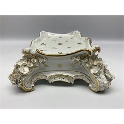 Late 19th century Meissen porcelain clock stand, of serpentine form with scrolling edges and applied flowers to the four scroll feet, heightened with gilt and painted with floral sprigs, with blue underglaze crossed swords mark beneath and incised number 572, H9cm W28cm D21.5cm, together with a further 19th century Continental porcelain stand, probably Furstenberg, of circular drum form with footed base, decorated in gilt with cornucopia and floral swags beneath gilt borders, with blue underglaze mark beneath, H15cm top D17cm