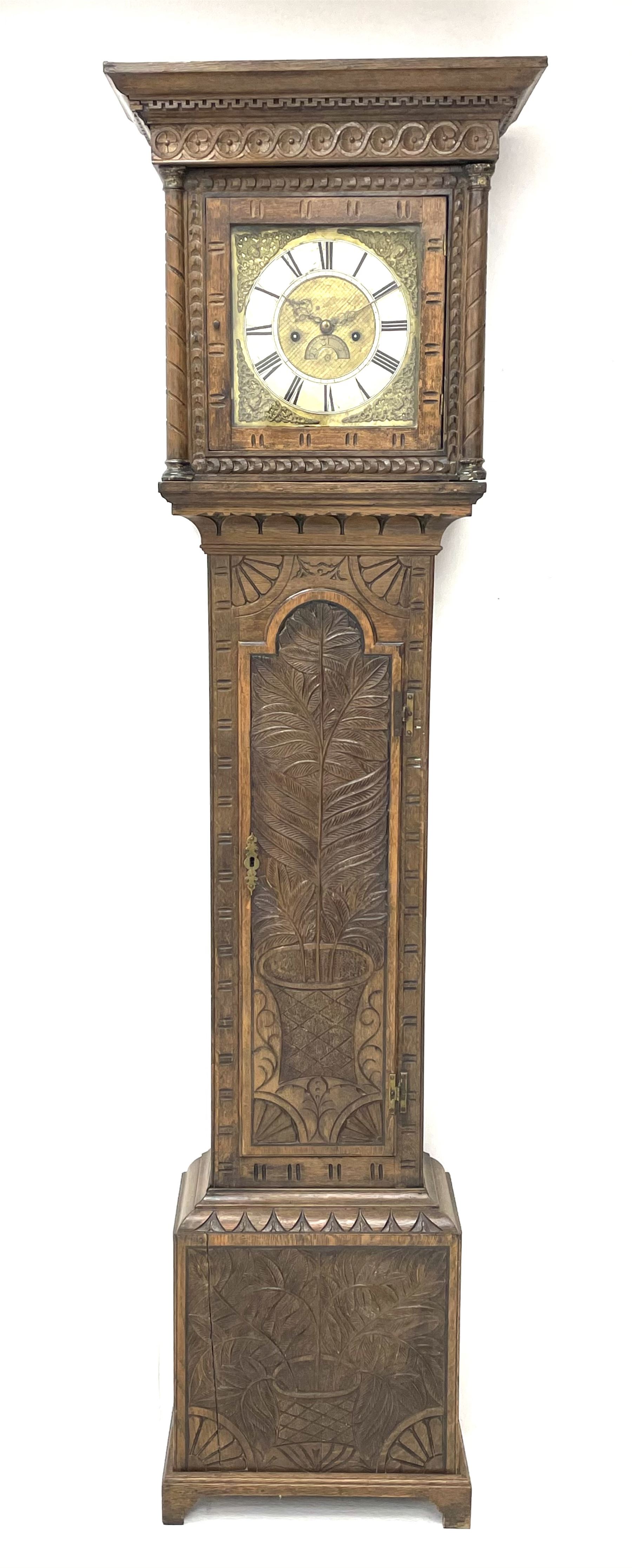 19th century carved oak longcase clock, projecting cornice over guilloche carved frieze, square brass dial with silvered Roman chapter ring and calendar aperture, decorated with ornate mask cast spandrels, the trunk door and base carved with potted plant, twin train driven eight day movement striking on bell