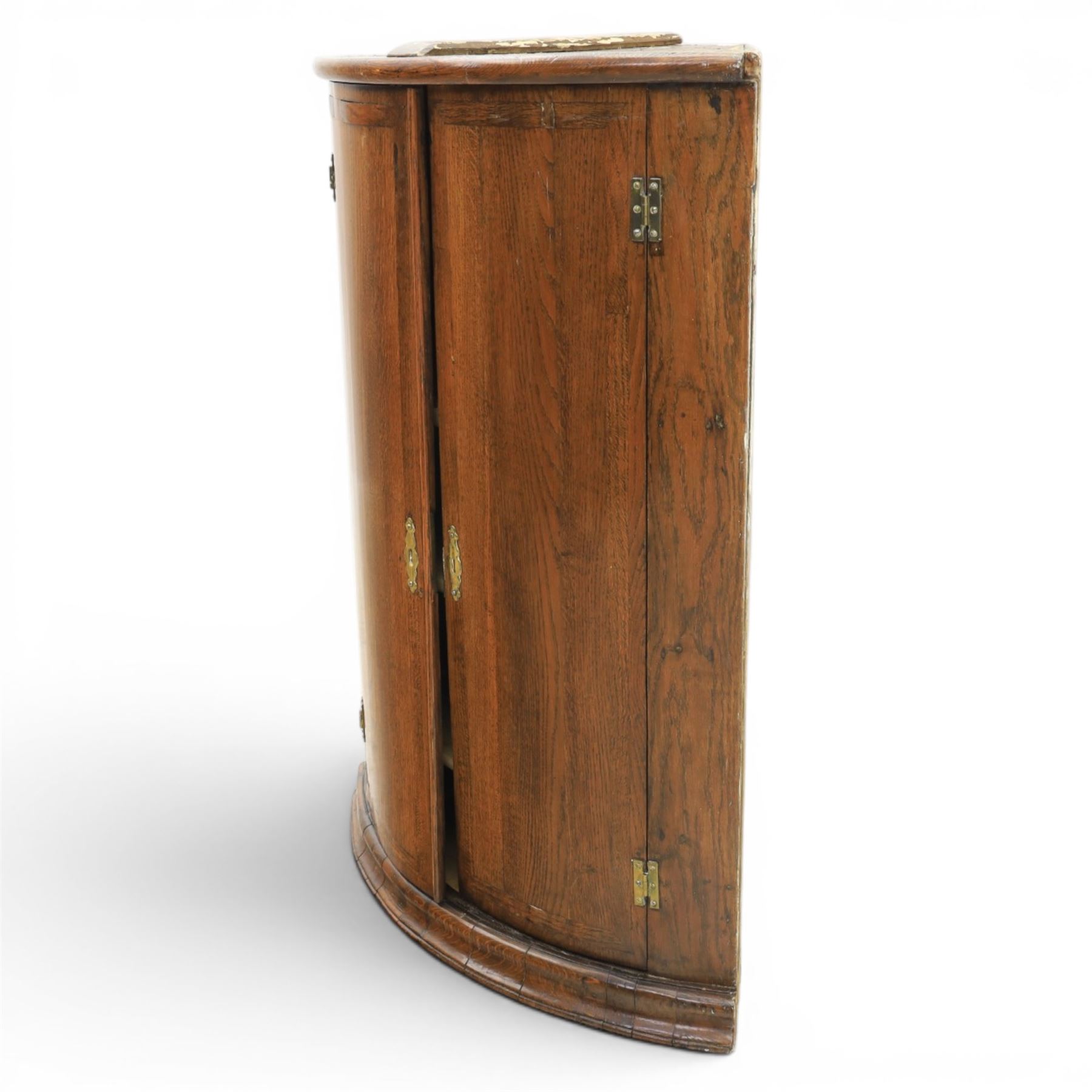 George III oak cylindrical corner cupboard, bow fronted doors revealing a painted shelved interior, brass escutcheons  and hinges, raised on a moulded plinth base