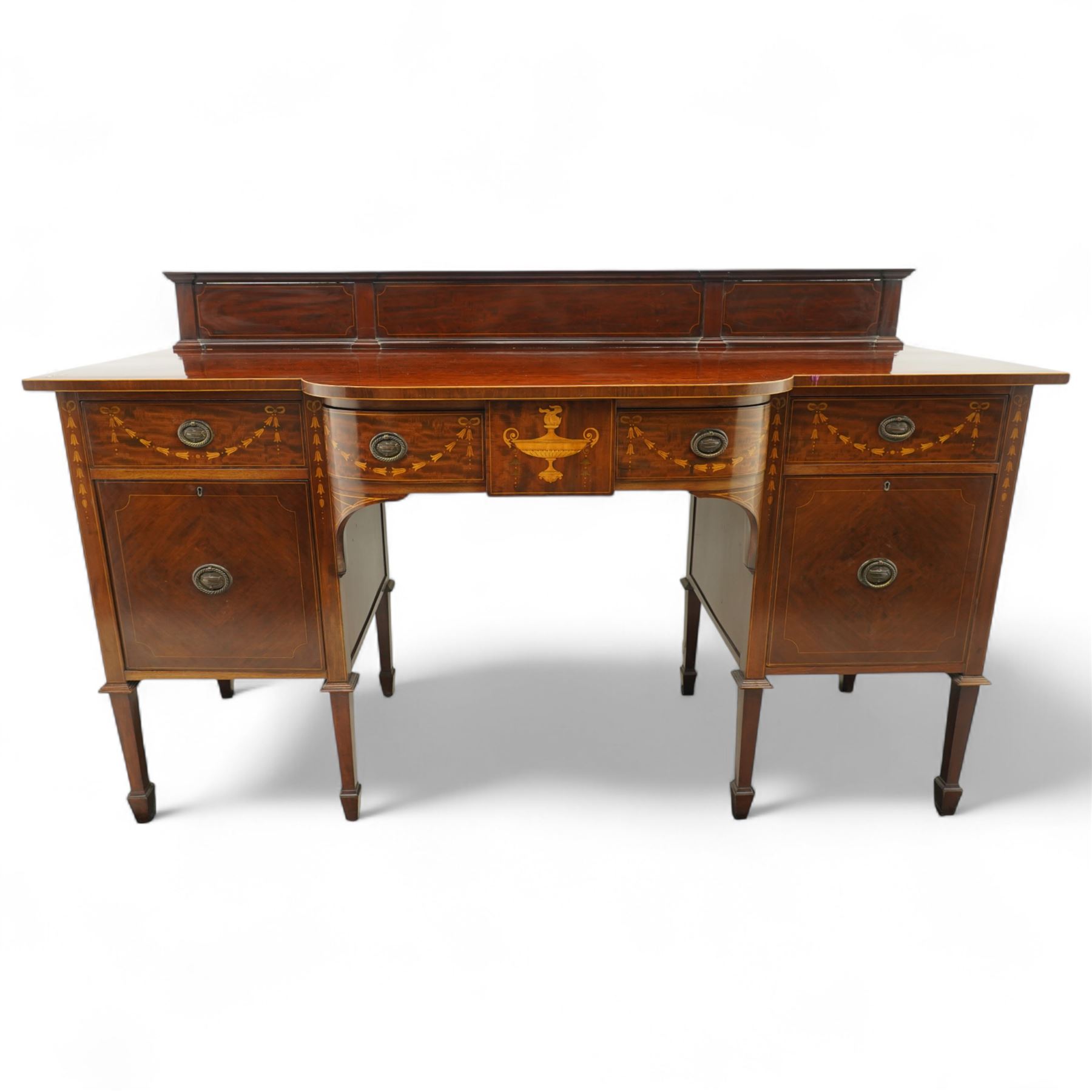 Edwardian Sheraton Revival inlaid plum pudding mahogany serpentine sideboard, rectangular top with raised gallery above bowed frieze drawer finely inlaid with central neoclassical urn motif, flanked by two drawers with swags and ribbon bows, above a cellarette drawer and cupboard, on square tapered supports with spade feet 