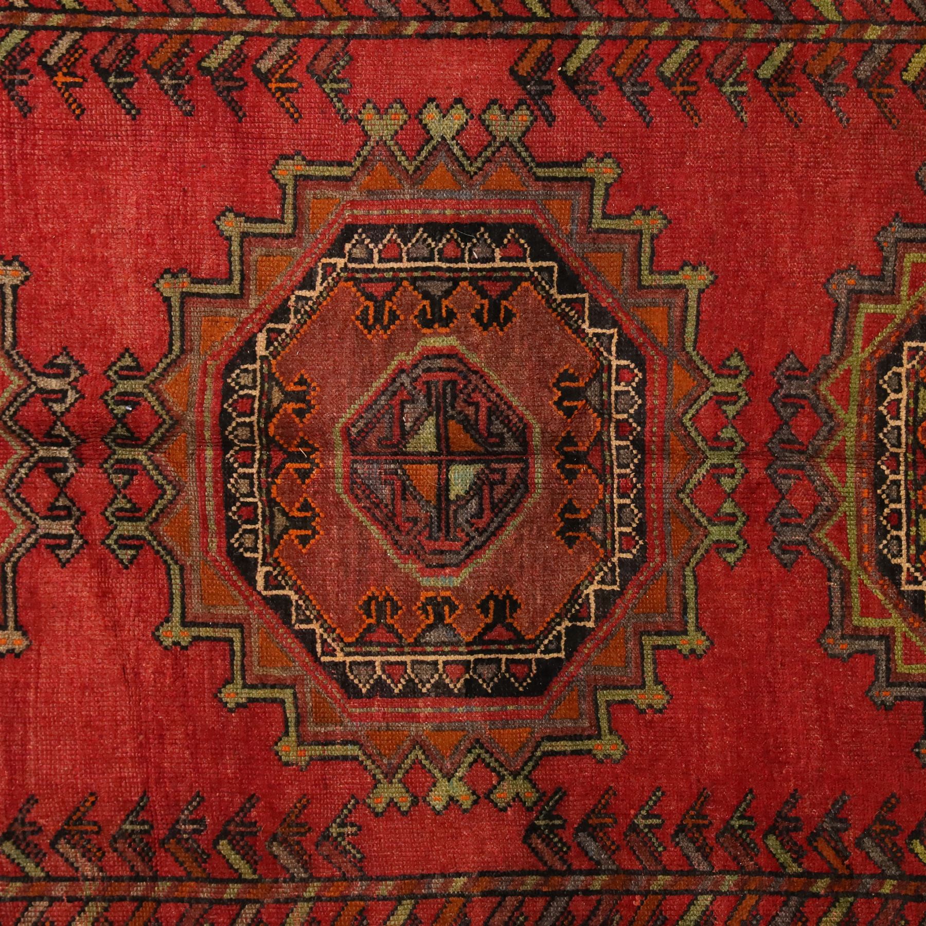 North East Persian Kurdish hand-knotted carpet, madder red field with three vertical and central stepped octagonal medallions enclosing angular geometric motifs with scattered tribal devices, framed by multiple narrow borders with latch-hook patterns, enclosing a bold main border of repeating geometric motifs, palette with indigo, dark brown and soft green tones, 