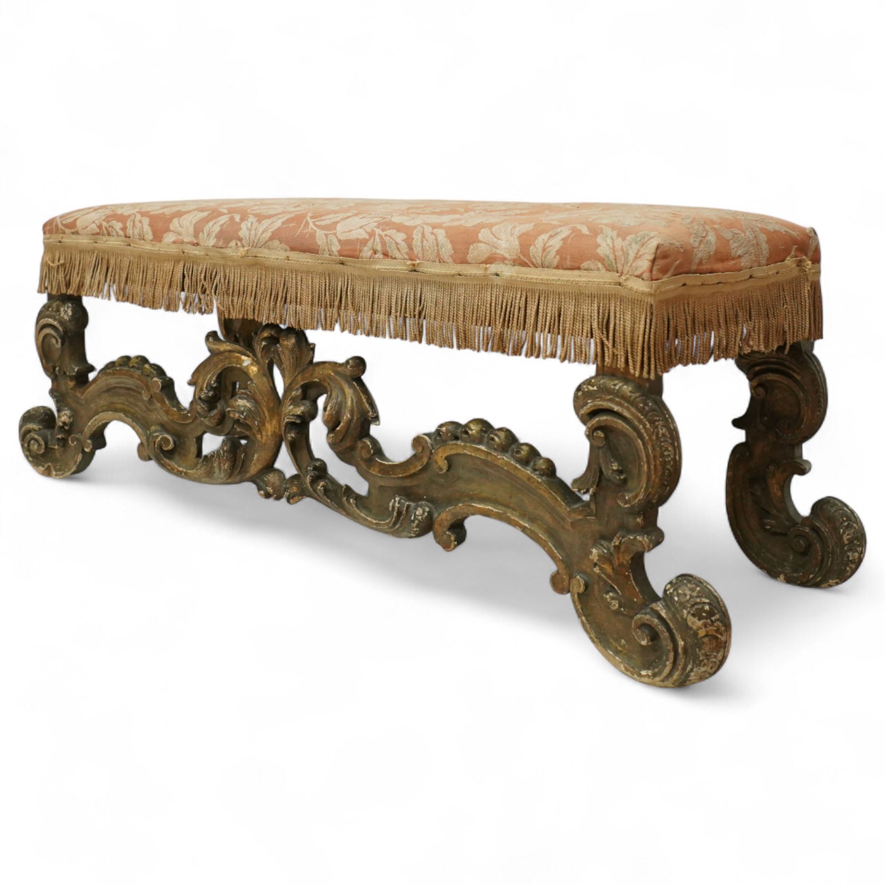 19th-century Rococo-style giltwood window seat, upholstered in floral-patterned coral-ground fabric with fringed border, the intricate scrolled supports united by pierced scrolling foliate stretchers with acanthus and C-scroll decoration